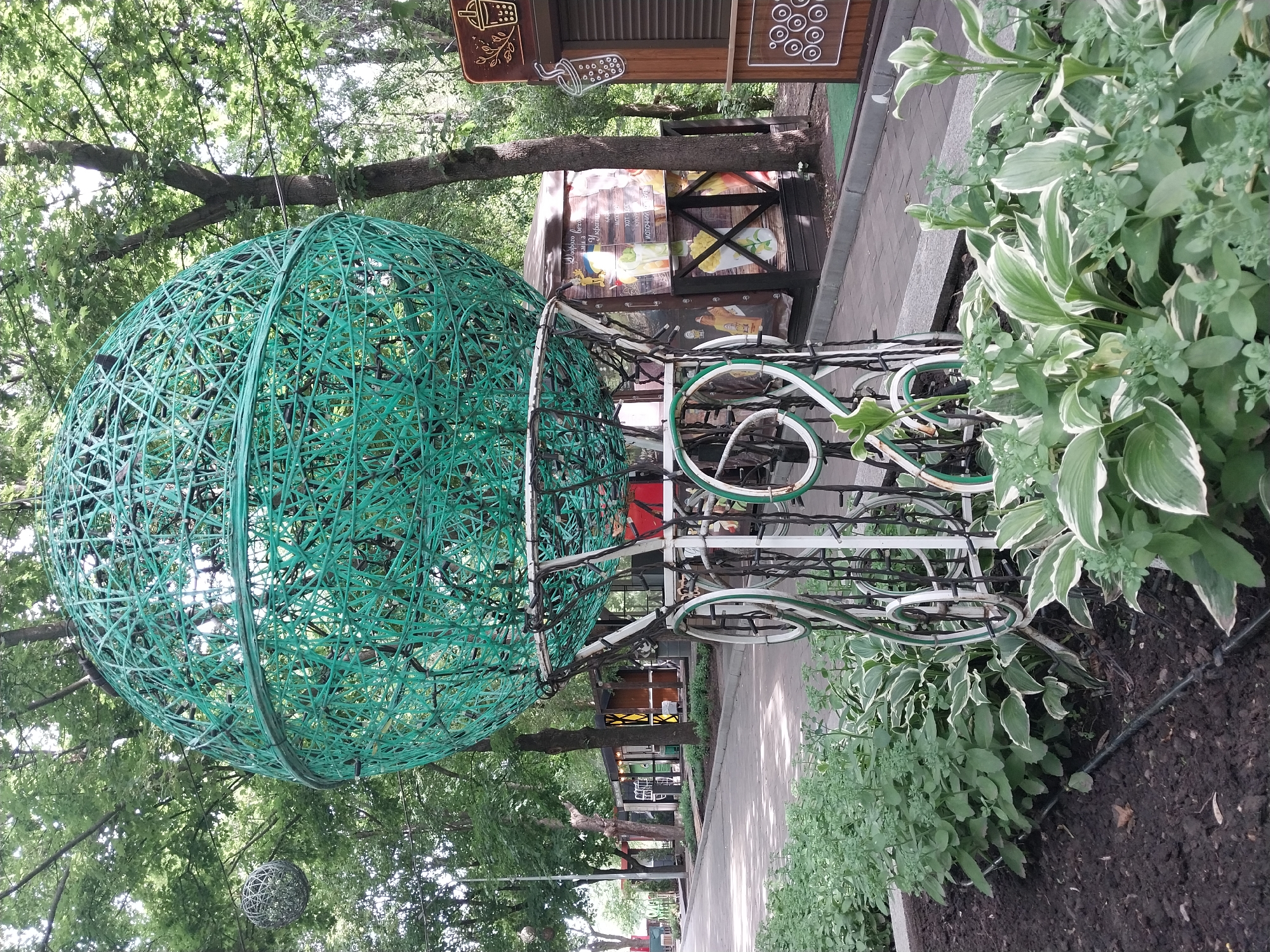 The image depicts an outdoor scene in a park in Dnipro, Ukraine, likely during daytime. A large, spherical sculpture, constructed from interwoven green material, dominates the foreground. This sculpture is elevated on a white, ornate metal stand that resembles a vintage carriage or hot air balloon basket, complete with decorative swirling elements and wheels.
Behind the sculpture, a paved walkway stretches into the distance, lined with trees and several small kiosks or stalls. These stalls appear to be for food or drink vendors, with signage and colorful graphics visible. One stall prominently features imagery of drinks and text in Ukrainian.
The foreground is bordered by a garden bed with various plants, including lush green foliage and some hostas with variegated leaves. The ground is covered in dark soil. The overall atmosphere suggests a recreational area, possibly a park or esplanade, designed for public enjoyment. The presence of trees and greenery indicates a natural setting within an urban environment.