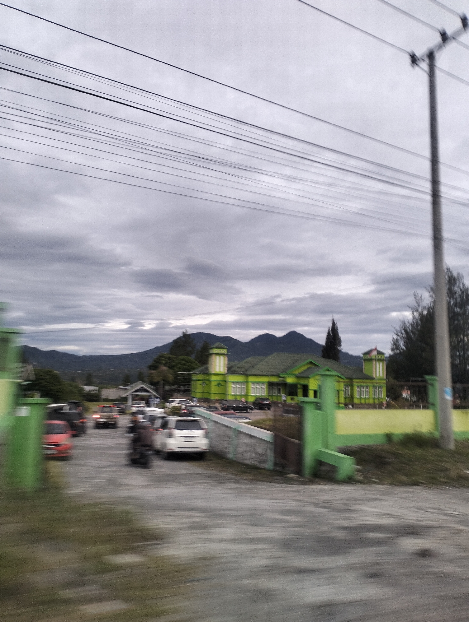 This media file captures a scene in Panteraya, Indonesia, likely during the day under overcast skies. The foreground is blurred due to motion, suggesting the image was taken from a moving vehicle. Several vehicles, including cars and a motorcycle, are visible in what appears to be a parking area or road.
In the middle ground, a distinctive green building with yellow accents stands prominently. It has a symmetrical design with multiple windows and a tower-like structure on the right, topped with a red and white flag. The building's architecture suggests it could be a public institution, a school, or perhaps a place of worship.
In the background, rolling hills and mountains are silhouetted against a cloudy sky. The overall atmosphere is somewhat muted due to the cloudy weather, but the bright green of the building provides a visual anchor. The presence of power lines overhead is a common sight in many populated areas and adds to the everyday urban or suburban setting. The motion blur in the foreground and the general lack of sharp detail prevent the identification of specific individuals or detailed emotional expressions.