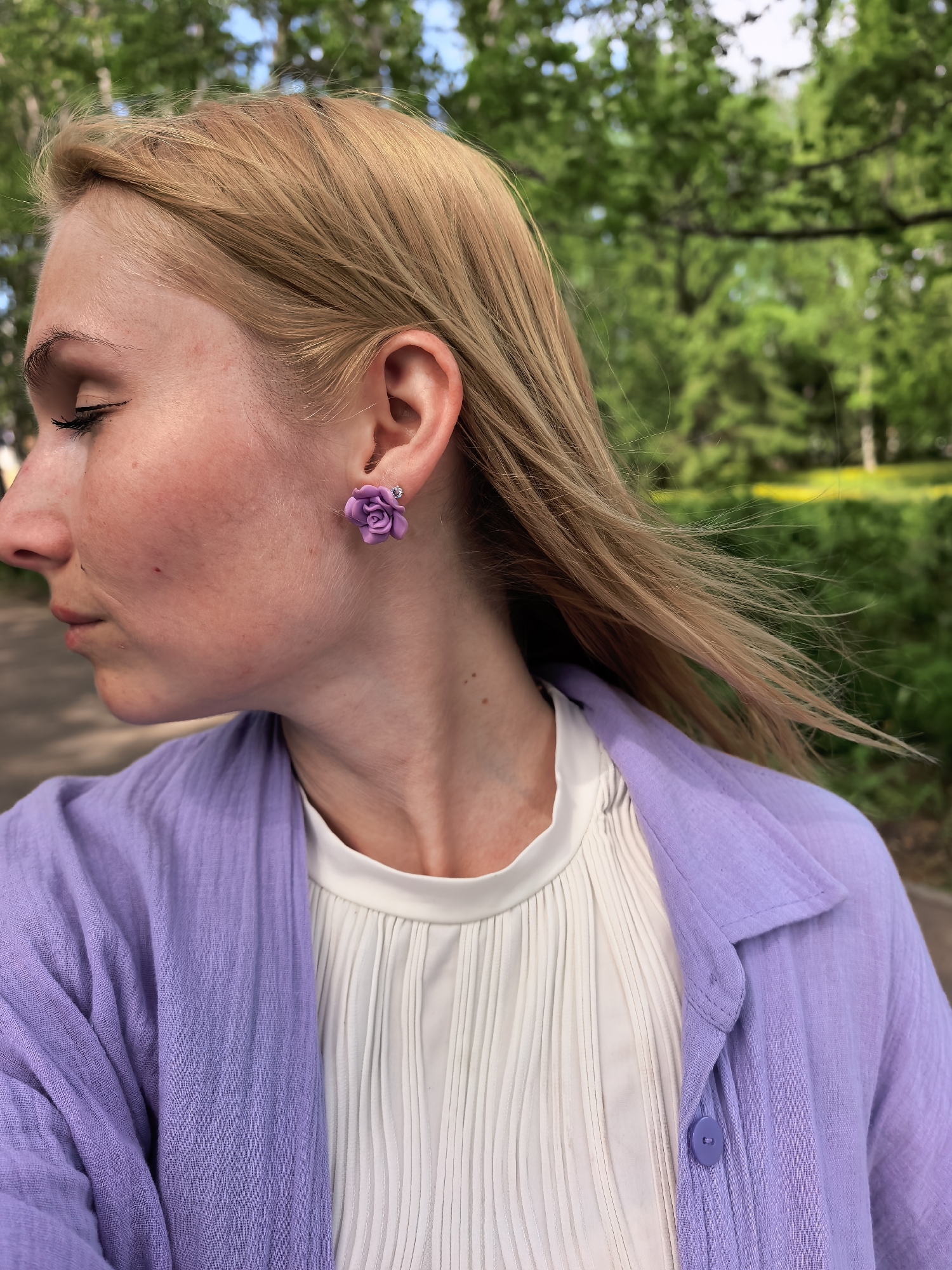A woman is seen in a medium shot, taken in what appears to be an outdoor park setting. The woman is turned in profile, facing left with her eyes closed or lowered. She has fair skin and light brown or blonde hair that is partially blown by a breeze. She is wearing a white pleated blouse with a round neckline, and a light purple or lavender button-down shirt or jacket. A distinctive feature is the large, purple flower-shaped earring on her visible ear. The background consists of green foliage, suggesting trees and shrubbery, blurred in the background, likely due to depth of field. The location is in the city of Sterlitamak, Russia.