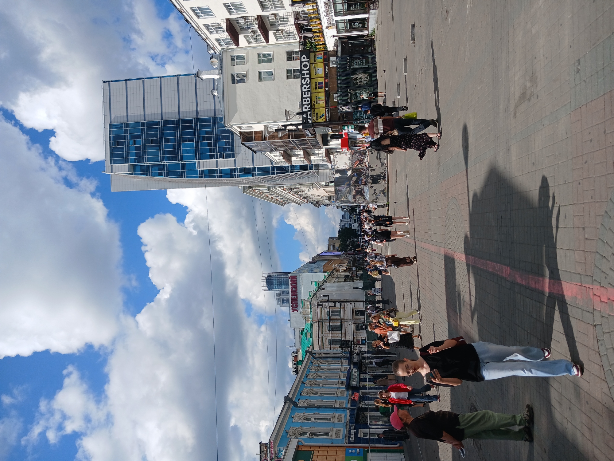 The image depicts a street scene in Yekaterinburg, Russia, on a partly cloudy day. The viewpoint is from a pedestrian's perspective, looking down a wide, paved street lined with buildings of varying architectural styles.
In the foreground, several people are walking on the sidewalk. A woman in a black top and light blue jeans is walking towards the camera, looking at her phone. To her left, a man in a green shirt and pants and a woman in a red jacket are standing. Further down the street, more pedestrians are visible, creating a sense of urban activity.
The buildings on either side of the street display a mix of historical and modern architecture. On the left, there are ornate, older buildings with blue facades and decorative white trim. On the right, a tall, modern glass-fronted building stands out, contrasted with adjacent multi-story buildings featuring balconies and window units. Some shops are visible, including one with a prominent "BARBERSHOP" sign.
The sky is a prominent feature, with large, white cumulus clouds scattered across a bright blue expanse. The sunlight casts shadows on the pavement, indicating it is likely daytime. The street is paved with interlocking bricks. In the background, more buildings and a distant tower can be seen, contributing to the cityscape. The overall impression is of a busy urban environment.