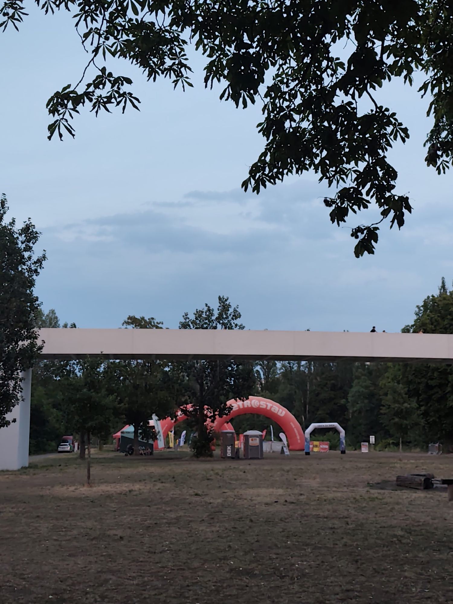 The image depicts an outdoor scene at dusk, likely in Prague, Czechia, with a prominent white pedestrian bridge spanning across the frame from left to right. In the background, several inflatable arches are visible, indicating an event or race. One large red inflatable arch displays text that reads "THESAUDA." Other smaller arches and tents are scattered across the field. Trees line the perimeter of the area, and the sky is overcast with soft clouds. A few people are visible on top of the pedestrian bridge, and a red vehicle is parked in the distance to the left. The ground is covered with dry grass.
