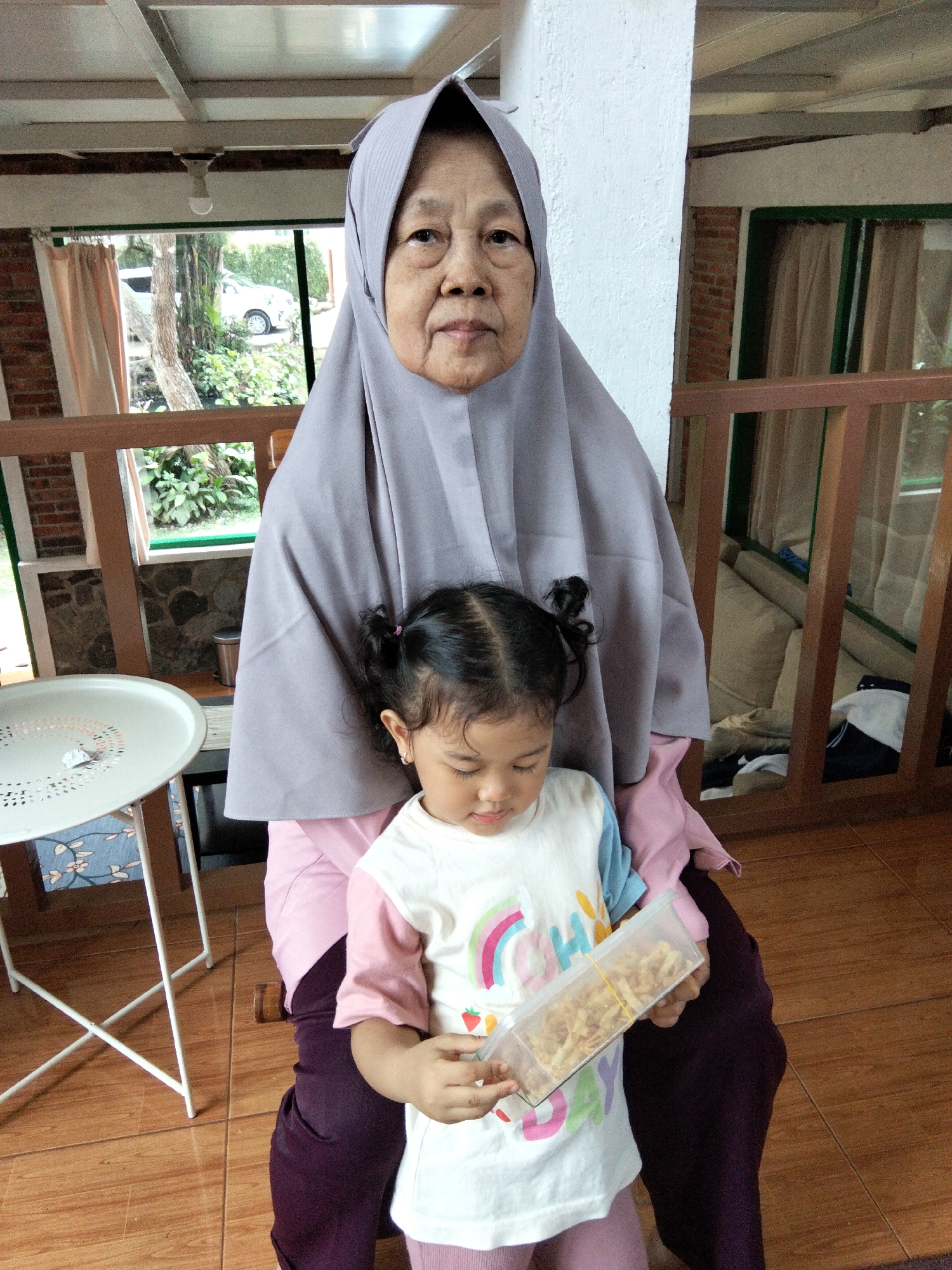 An elderly Indonesian woman wearing a light purple hijab and a pink top is seated, holding a young Indonesian girl on her lap. The girl, with her hair in two pigtails, wears a white t-shirt with colorful graphics and is holding a transparent plastic container of food. They are indoors, on a wooden floor, with a white table and chairs to the left. In the background, there is a wooden railing, a window showing greenery and a parked car, and furniture. The overall setting suggests a relaxed, domestic environment, possibly a cafe or a home in Cisarua, Indonesia.