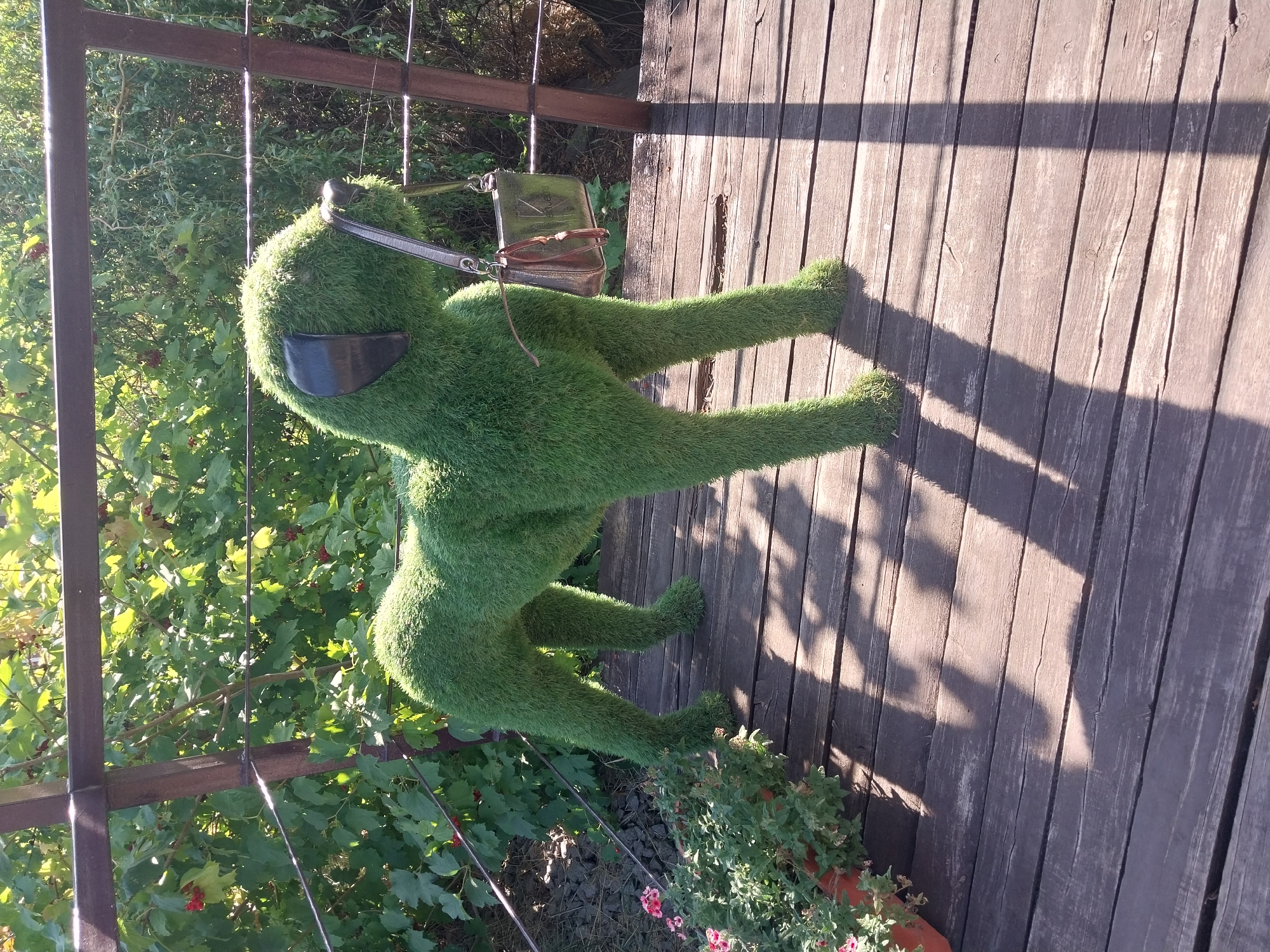 In Petrykivka, Ukraine, a topiary dog sculpture made of artificial grass stands on a wooden deck. The dog is positioned mid-stride, with its mouth holding a metallic purse by the strap. The sculpture is detailed, resembling a real dog, with black ears and a snout. Behind the dog, a metal railing is visible, with lush green foliage and some red berries growing beyond it. In the foreground to the left, a potted plant with small pink flowers adds a touch of color to the scene. The wooden deck exhibits a weathered appearance with visible grain and some cracks, and is partially covered by shadows cast by the railing and the dog sculpture, suggesting it is daytime.