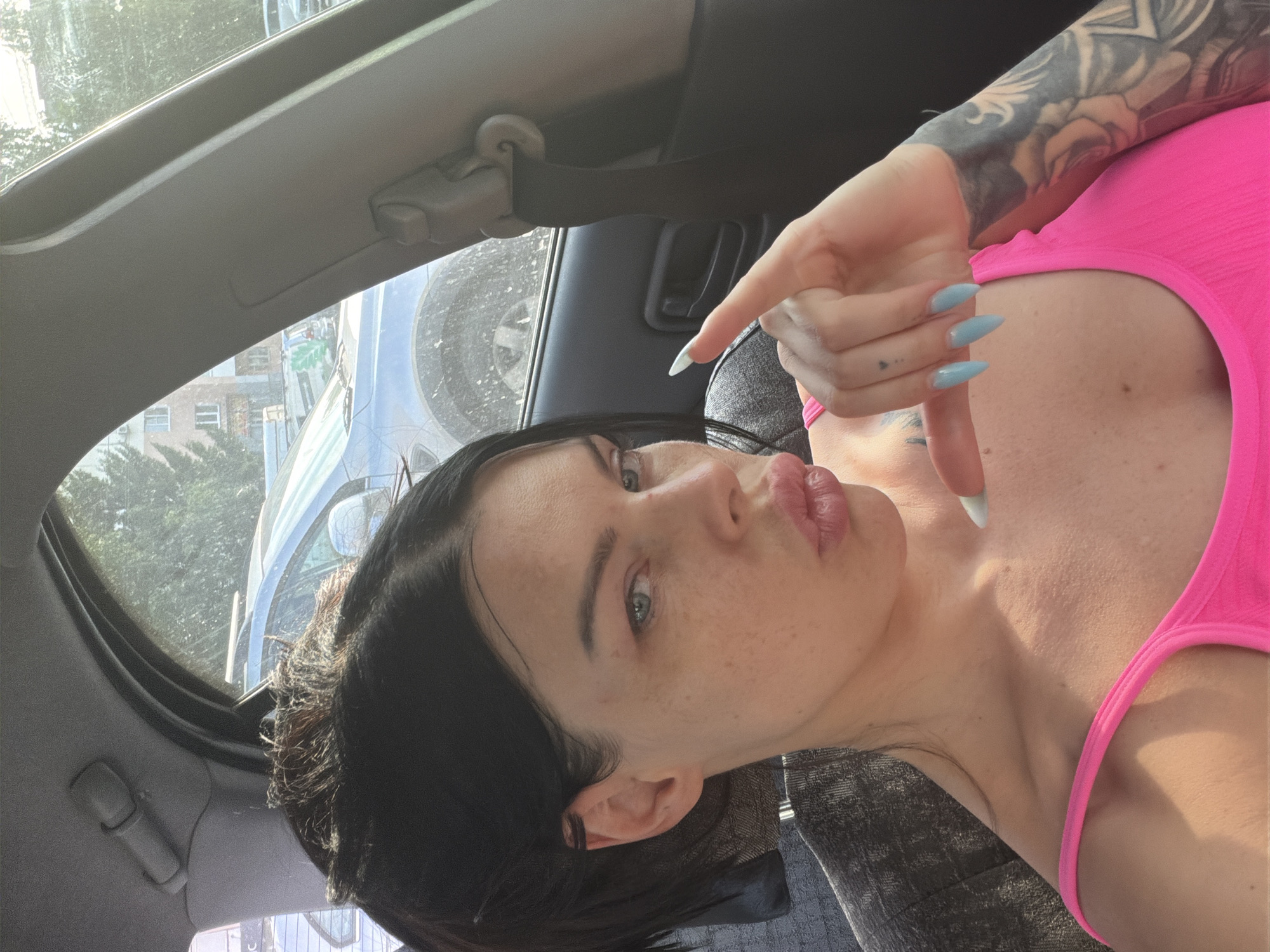 A woman with dark hair pulled back is inside a car. She is wearing a bright pink top and has light blue fingernails. The woman is looking at the camera and has her lips pursed. Her right hand is raised, with her index finger pointing forward. Visible tattoos are on her right arm. Outside the car, another vehicle and a building can be seen. The scene appears to be in an urban environment.