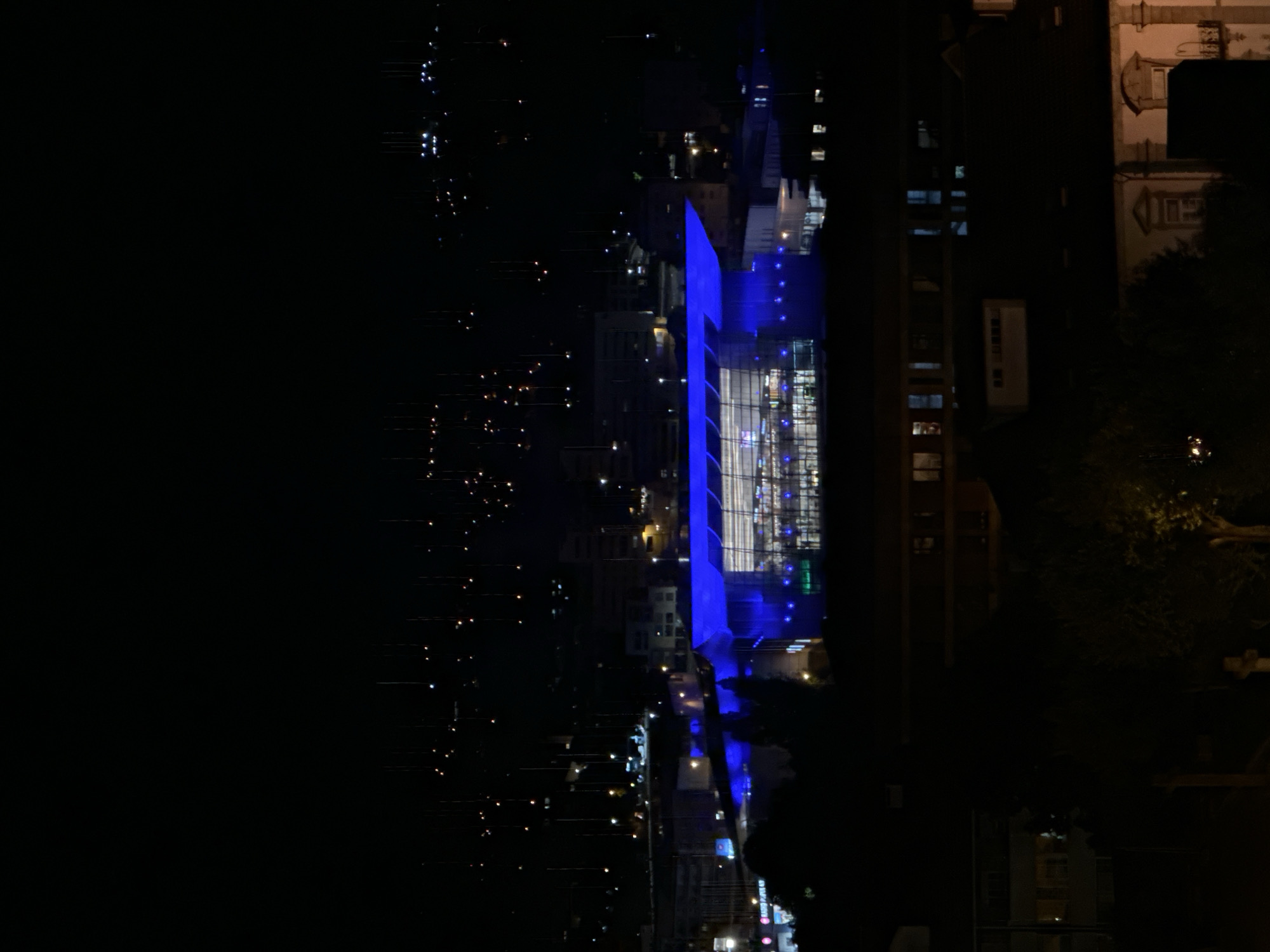 The image captures a nighttime view of Vila Real, Portugal. The dominant feature is a modern architectural structure, possibly a bridge or large building, brightly illuminated with a striking blue light. This blue light casts a reflection onto a surface below, suggesting the presence of water, such as a river or lake.
To the left of the prominent blue structure, the dark expanse of the night sky or distant landscape is dotted with numerous small, scattered lights of varying colors and intensities. These lights likely belong to other buildings, streetlights, or parts of the city extending into the distance, hinting at an urban or semi-urban environment.
On the right side of the frame, contrasting with the modern blue illumination, are parts of older buildings with more traditional architectural styles. One building on the far right has a light-colored facade, visible window frames, and is lit with a warmer, yellowish light. Dark, silhouetted shapes, likely trees, are also visible on this side, adding to the nocturnal landscape.
The scene is devoid of people or discernible activity, focusing instead on the interplay of artificial light and architecture against the dark night. The time of day is clearly night, with no natural light source visible, and the weather appears clear. No visible text is present in the image.