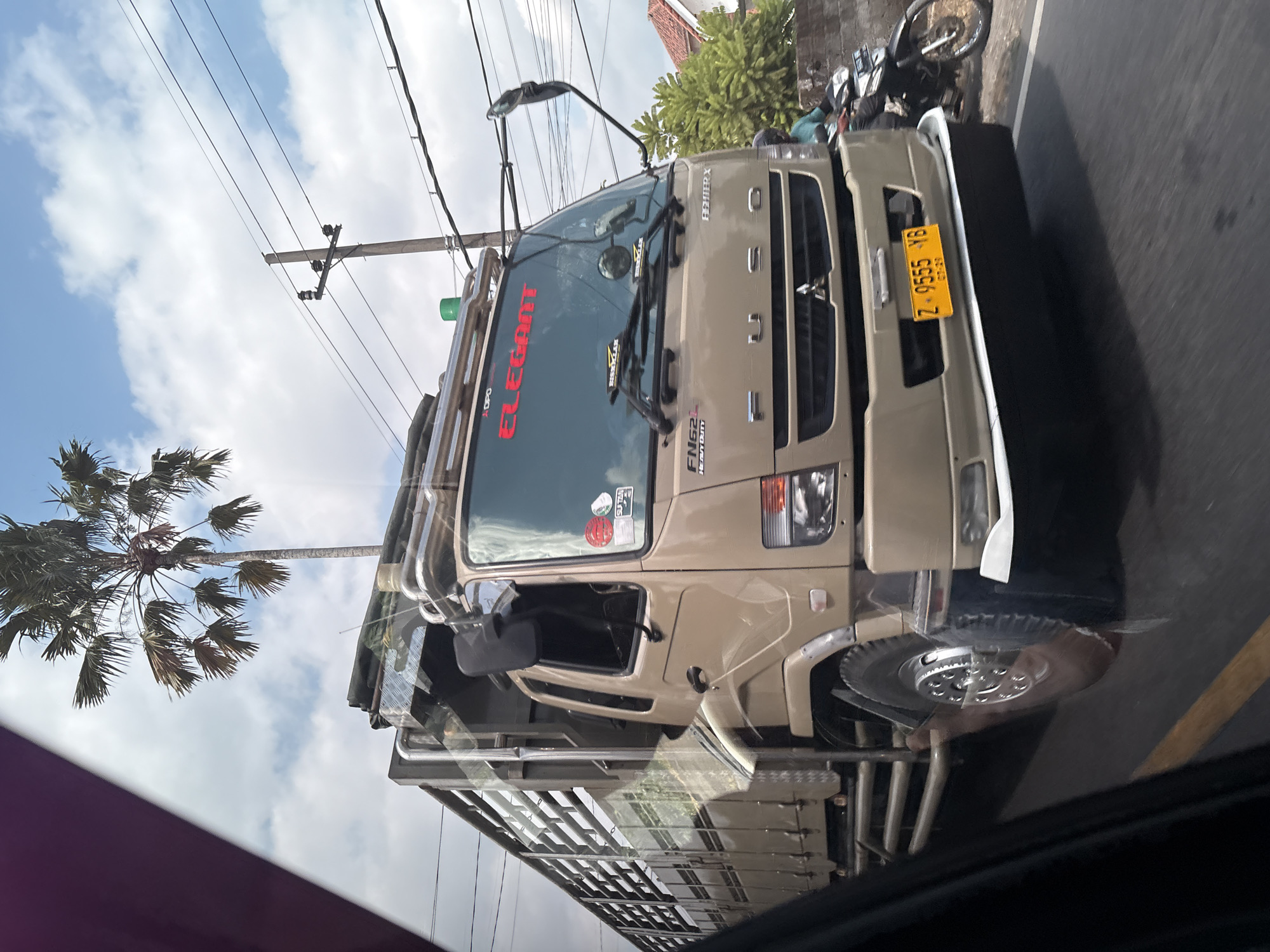 The image shows a tan FUSO truck with the word "ELEGaNT" written in red lettering across the windshield. The truck is driving on a road, with a motorcycle passing on the right. There is a palm tree and utility poles in the background, indicating an outdoor, possibly urban or suburban setting. The license plate of the truck is visible and reads "Z-9555-YB". The sky is partly cloudy. The image appears to be taken from inside another vehicle.