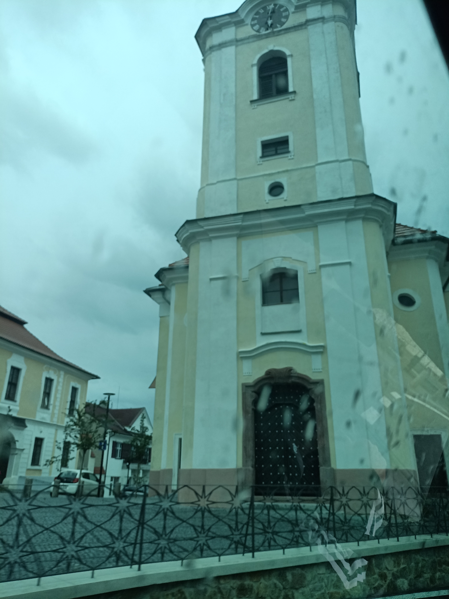 The image shows a church tower in Dalovy, Czechia, on an overcast day. The tower is tall and slender, with a clock near the top. It is painted in a pale yellow and white color scheme. There are several windows visible on the tower, some arched and some rectangular. At the base of the tower, there is a large, dark-colored door with decorative metalwork. In front of the church is a paved courtyard with a decorative metal fence. To the left of the church, there are other buildings, including a building with a red-tiled roof and several windows. A white car is parked in the courtyard. The overall scene is one of a quiet, historical town square.