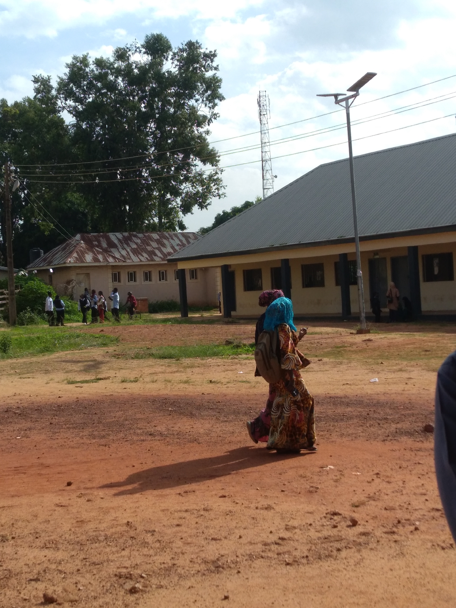 A group of people are walking on a dirt road in front of buildings. The buildings appear to be low-rise structures with light-colored walls and dark roofs. One building has a corrugated metal roof. There are trees and a cell tower in the background. The sky is partly cloudy. Two women in the foreground are wearing brightly colored clothing and headscarves. One is carrying a backpack. The scene is set in Jos, Nigeria.