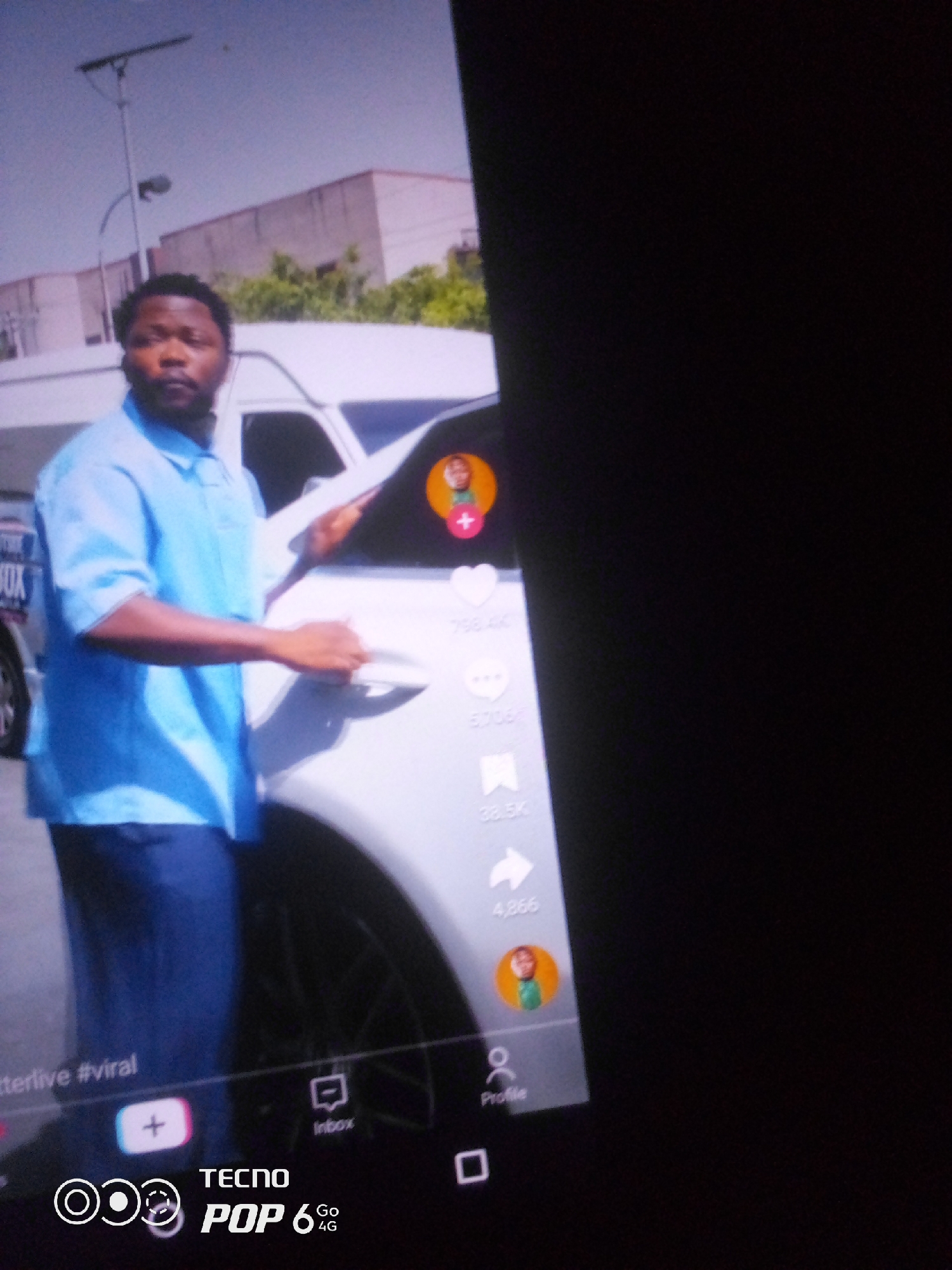 The image shows a man in a light blue short-sleeved collared shirt and dark pants standing next to a white vehicle. He has dark hair and a beard. He is touching the door handle of the vehicle. On the side of the vehicle, there are social media icons and engagement numbers, suggesting this may be a screenshot from a social media app. The background shows a building with a cloudy sky above, indicative of an outdoor urban setting. There is also a street light visible. The lower part of the image displays branding for "TECNO POP 6 4G". The overall scene appears to be a casual outdoor setting in a city. There is no specific information in the image to confirm the location in Howayi, Nigeria, but the presence of a vehicle, buildings, and urban infrastructure suggests a city environment.