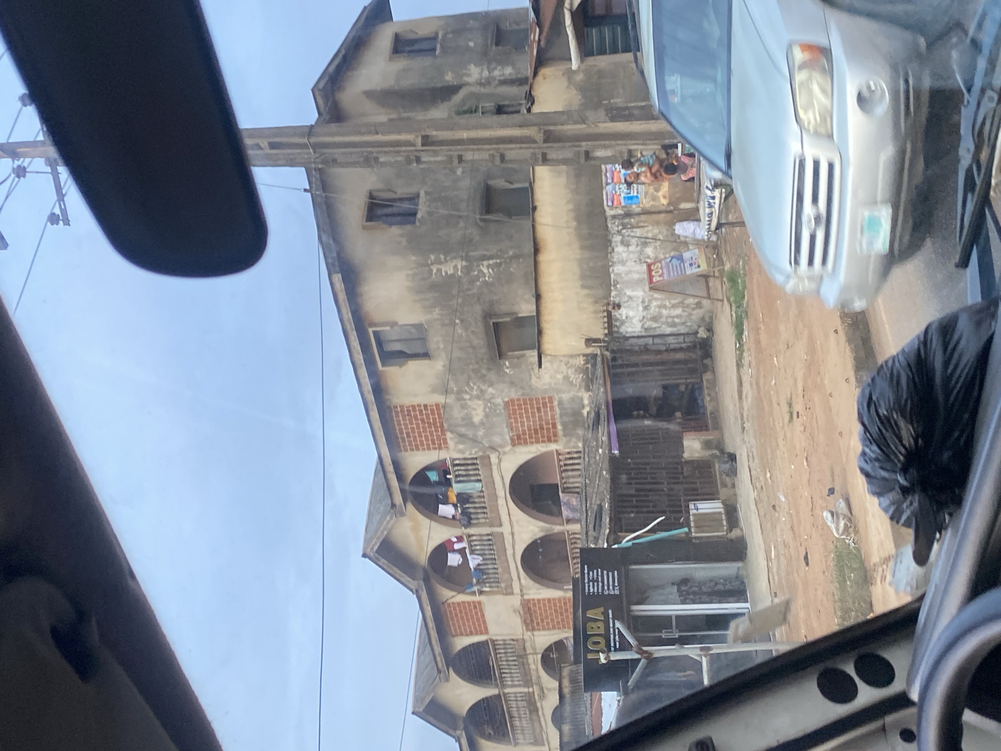 The image depicts a street scene in Ikorodu, Nigeria, characterized by a multi-story building with arched balconies adorned with laundry. A silver SUV is parked in the foreground, partially obscuring a dirt road and a few scattered items on the ground. Adjacent to the building is a storefront with a sign that reads "JOBA." The sky is overcast, suggesting a cloudy day.