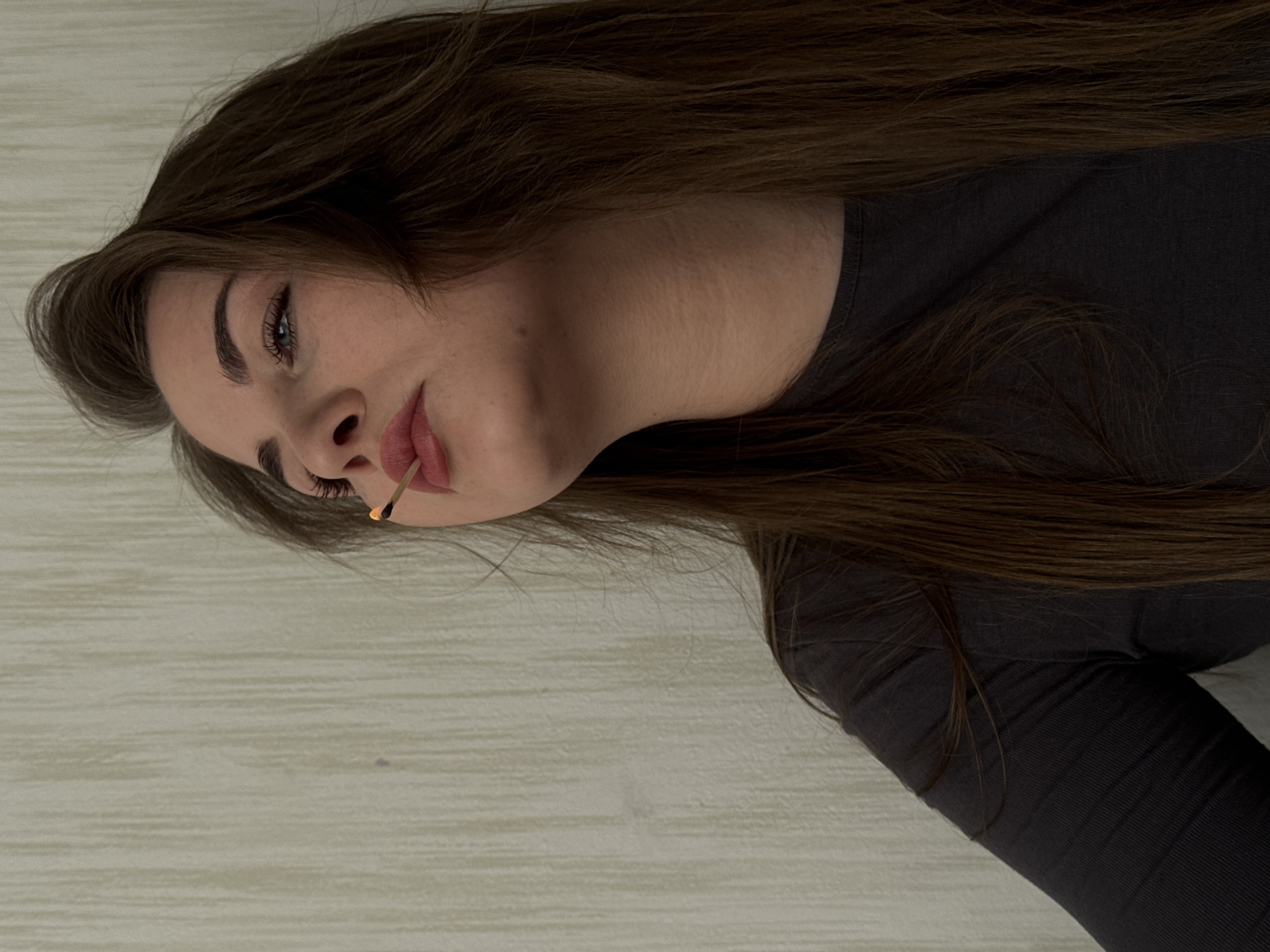 A woman with long brown hair is lying on a light-colored surface, possibly a bed or floor, with her head facing the camera. She is wearing a dark gray, long-sleeved shirt. She is holding a lit match in her mouth. The background appears to be a plain wall. There are no discernible details that point to the city of Chaikovsky, Russia. The information about the location is not evident from the image.