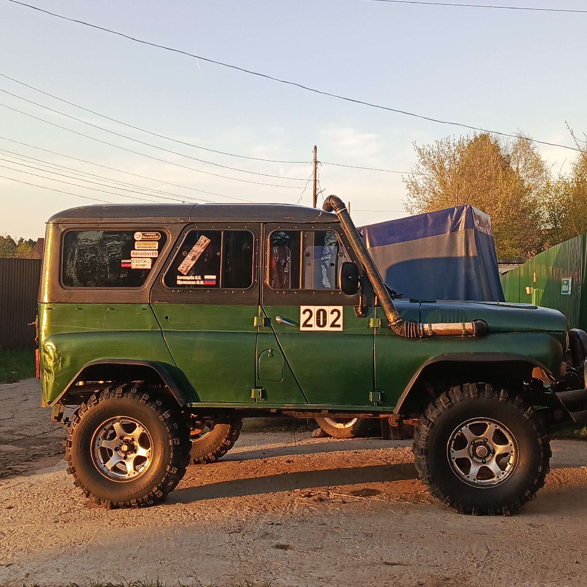 A green and black UAZ-469 off-road vehicle is parked on a dirt road. The vehicle has a snorkel intake on the passenger side, and the number "202" is displayed on the door. The vehicle's tires are large and have an aggressive tread pattern. Several stickers are visible on the rear window. In the background, a dark blue and black tarp covers an object. There's also a green fence and trees, along with power lines against a light blue sky. This image was taken in the city of Chaikovsky, Russia.