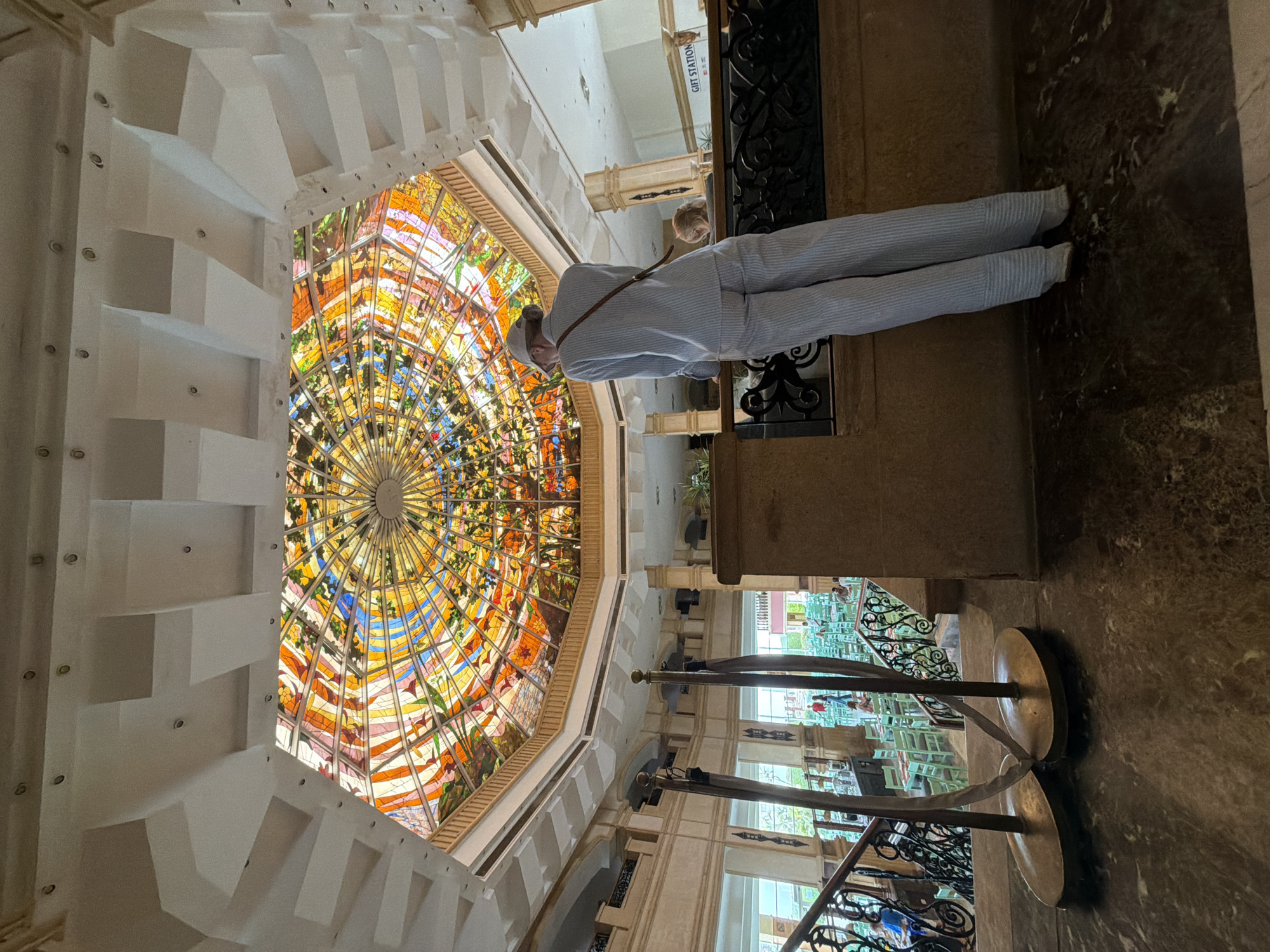 The image shows the interior of a building with a large, ornate stained glass dome. The dome features a vibrant circular design with various colors and patterns, illuminated by natural light. A person wearing light-colored striped clothing stands near a railing, looking up at the dome. The railing is decorated with intricate metalwork. A floor lamp with a brass base is also visible. The building's architecture includes white walls with stepped features. The location appears to be a high-end shopping mall with a gift station visible in the background, based on architectural style. Given the architecture and possible presence of a gift station, the location is most likely at the Il Mercato Shopping mall in Sharm El Sheikh, Egypt.