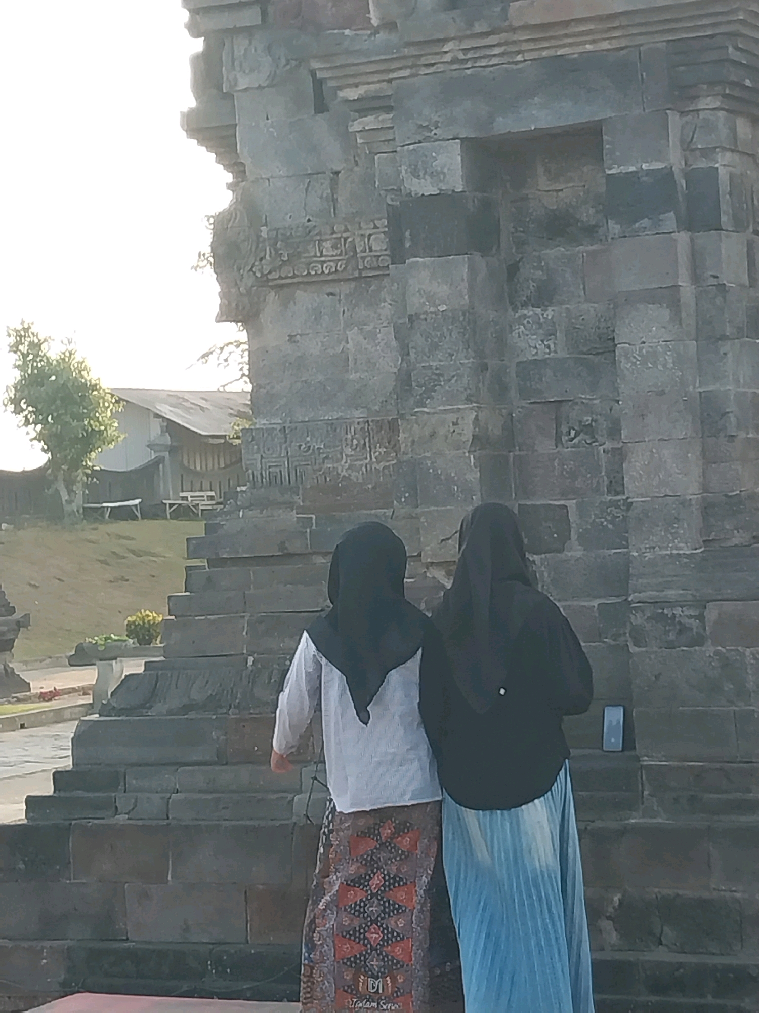 Two women, both wearing headscarves and long skirts, stand facing away from the camera. The woman on the left wears a white long-sleeved shirt and a patterned skirt. The woman on the right wears a black long-sleeved shirt and a light blue pleated skirt. They are positioned on stone steps in front of a large, weathered stone structure, possibly an ancient temple. The structure is made of large, rectangular blocks and shows signs of age and wear. Behind the women, to the left, is a grassy slope with a few trees and a building with a sloped roof. The sky is bright and hazy, suggesting it might be early morning or late afternoon. A small, rectangular electronic device is visible on the stone steps to the right of the woman in black. The scene appears to be outdoors in a historical or archaeological site.