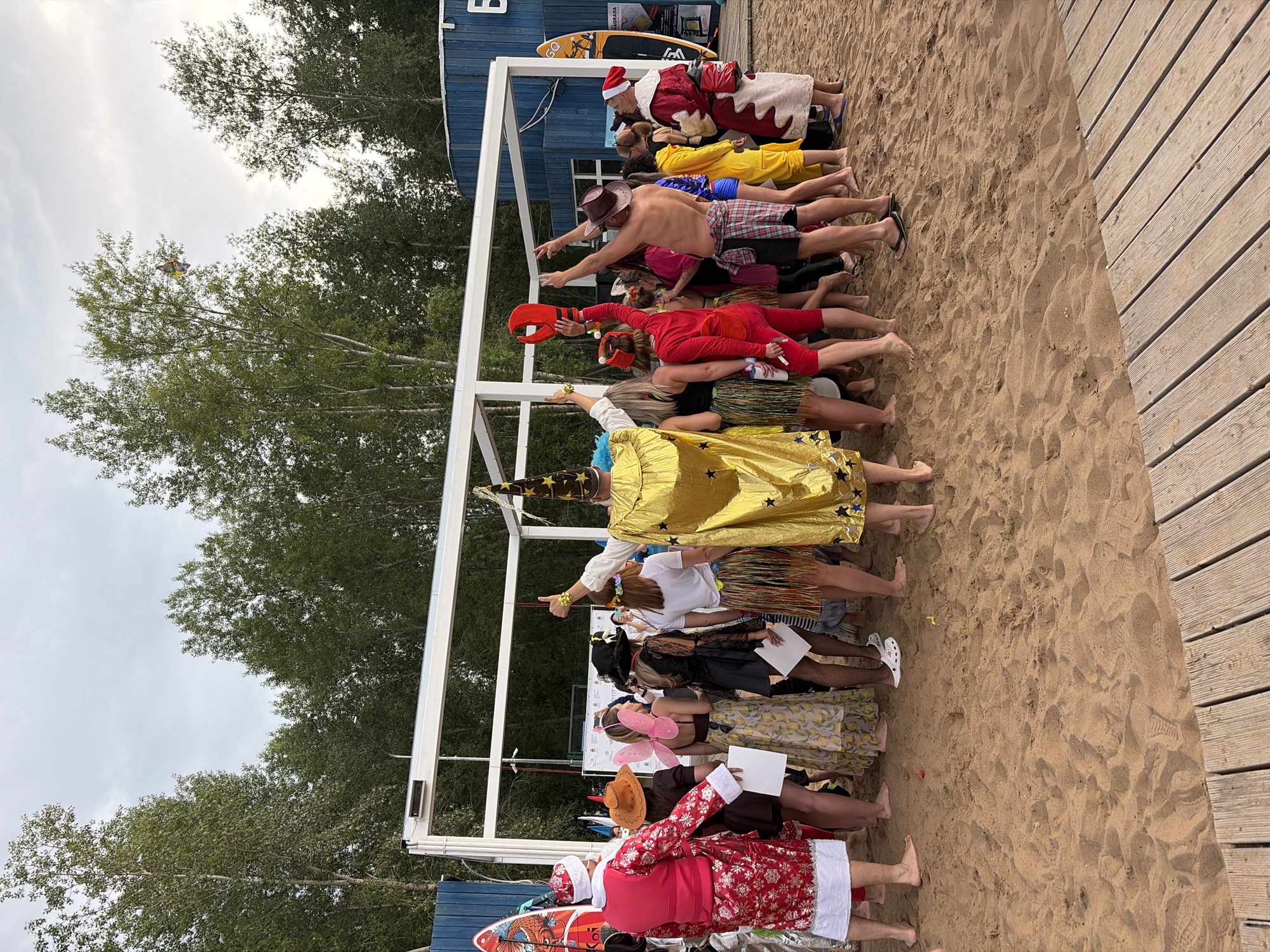 A group of people are dressed in costumes and standing in a line on a sandy beach. The person at the front is dressed as Santa Claus. Behind Santa, there are people in costumes resembling a lobster, a cowboy, a wizard, and a fairy. The scene appears to be an outdoor event, possibly a competition or a themed party. The sky is overcast, suggesting a cloudy day. In the background, there is a blue building and trees. A wooden boardwalk is visible on the right side of the image. Some of the people are holding white papers, which might be scorecards or instructions. The overall mood seems festive and playful. There is some text visible on a surfboard in the background, but it is not legible.
