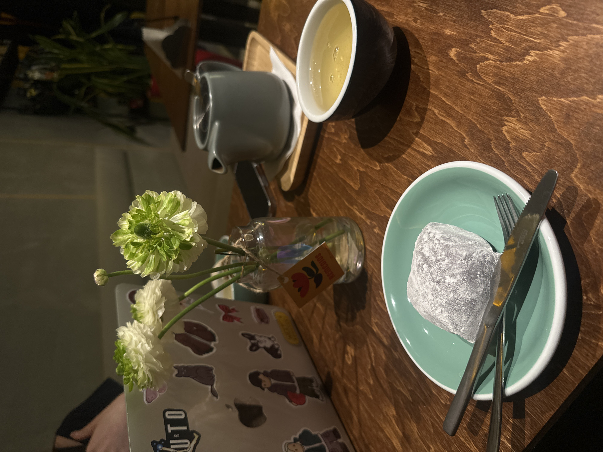 A still-life photograph captures a table setting, likely in a cafe or restaurant. The scene includes a laptop adorned with stickers, a small vase with white and green flowers, a light green plate with a dark gray mochi, and a knife and fork placed on top. A teapot and a cup of tea complete the setting. The background is partially obscured, but the presence of a seated person suggests an interior location. The location of the photo is not provided.