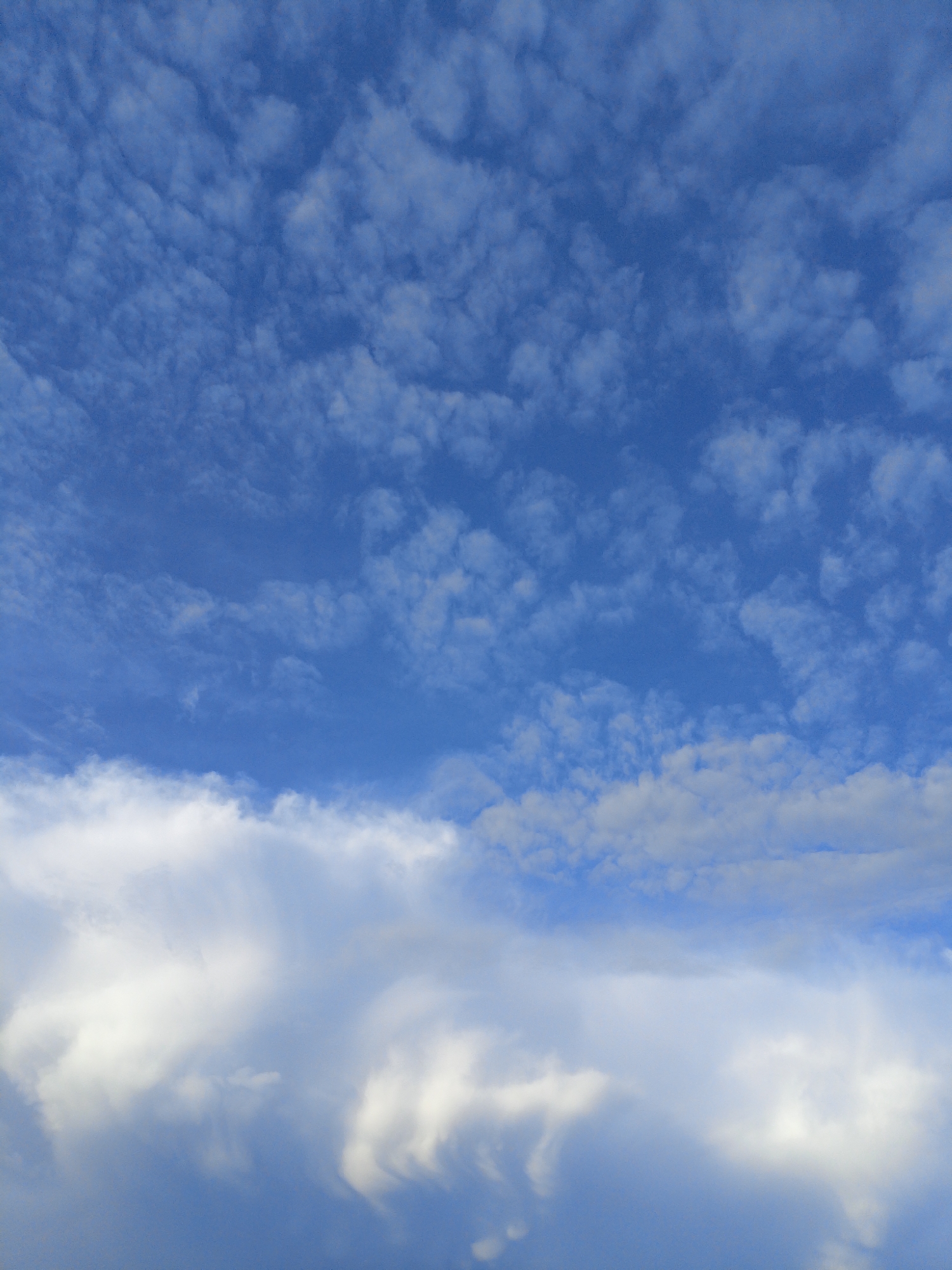 The image is a medium-angle shot of a blue sky filled with clouds. The clouds are varied, with some appearing as wispy, scattered cirrus clouds in the upper portion of the frame, and others as thicker, cumulus clouds in the lower half. The lighting suggests it is daytime, possibly late afternoon given the slightly muted tones in some of the cloud formations. There are no people, man-made structures, or discernible landmarks visible in the image. The scene is entirely focused on the atmospheric conditions. The location is unknown.