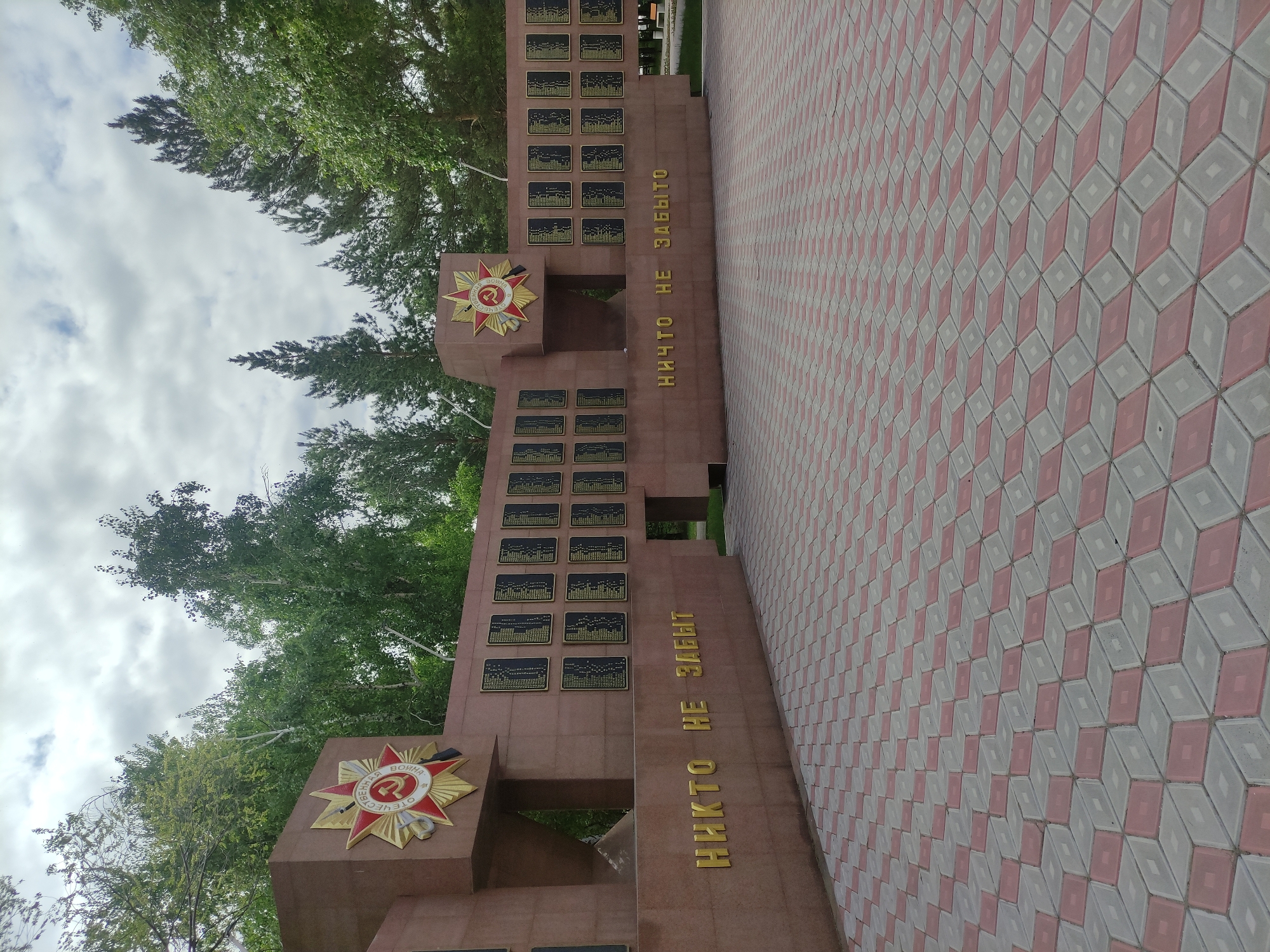 The image shows a memorial in Pavlodar, Kazakhstan, with a red star emblem and inscribed text in Russian: "НИКТО НЕ ЗАБЫТ, НИЧТО НЕ ЗАБЫТО" ("No one is forgotten, nothing is forgotten"). The memorial is constructed from brown stone or concrete and features multiple plaques. A patterned brick path leads up to it. The sky is cloudy and trees are visible in the background.