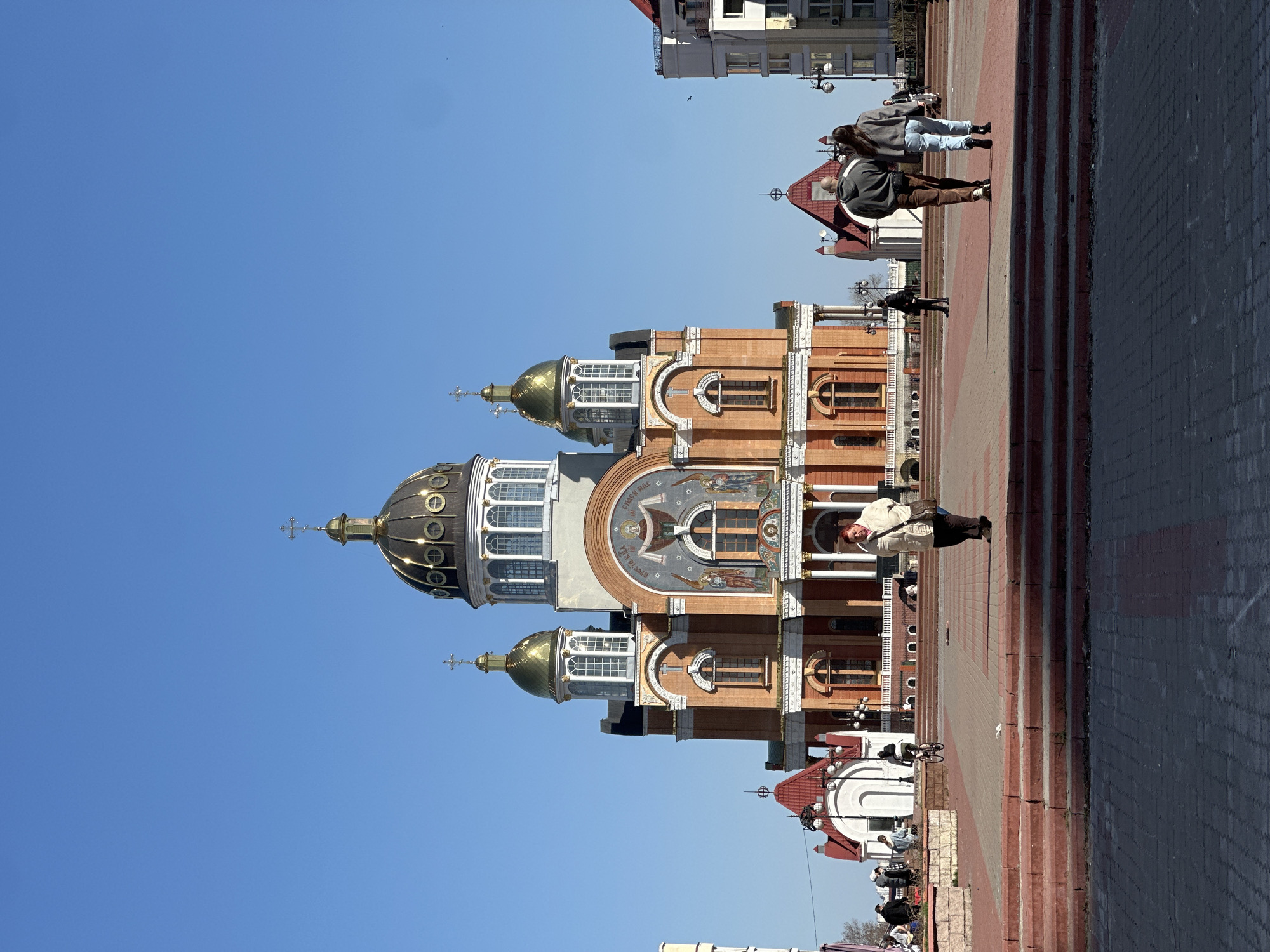 The image depicts the Patriarchal Cathedral of the Resurrection of Christ, located in Kyiv, Ukraine. The cathedral has a brick facade with ornate detailing and multiple golden domes topped with crosses. The sky is clear and blue.
Several people are visible in the foreground walking on a brick-paved area in front of the cathedral. One individual is wearing a light-colored jacket and carrying a bag, while another is dressed in darker clothing. The people appear to be interacting with the environment by strolling around the public square.
Buildings are visible in the background behind the cathedral. Street lights, red rooftops, and trees are also observed.