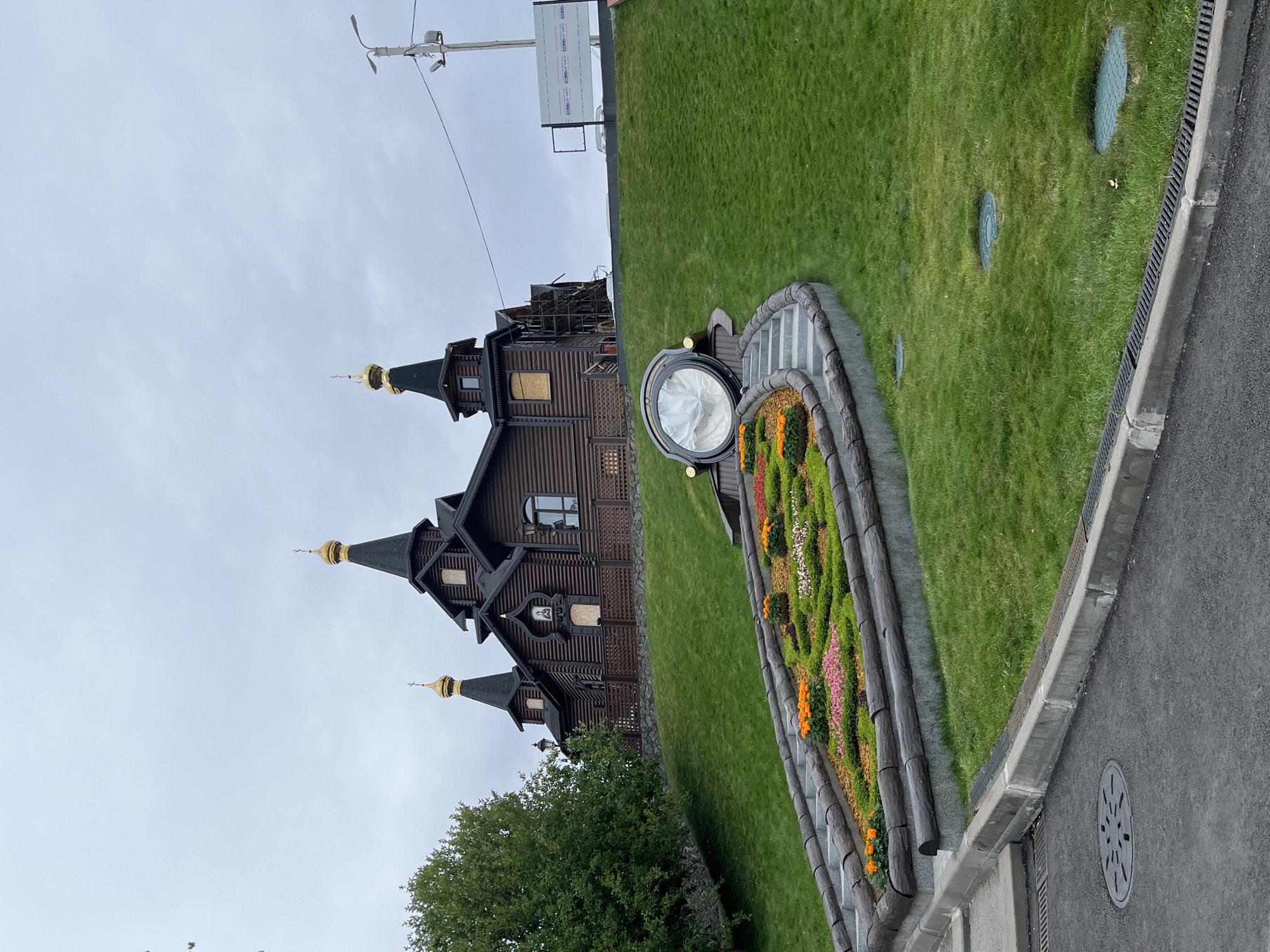 A brown, wooden, domed church with golden cupolas stands on a grassy hill under a cloudy sky. In front of the church, a circular flower bed filled with various colorful flowers and surrounded by wooden logs is visible. A large, circular glass structure is located at the top of the flower bed. Steps lead up to the flower bed and the church. To the right of the church, a tall utility pole with wires is present, along with a white construction fence. The ground in front of the hill is paved with asphalt and has a manhole cover. The scene is located in Kharkiv, Ukraine.
