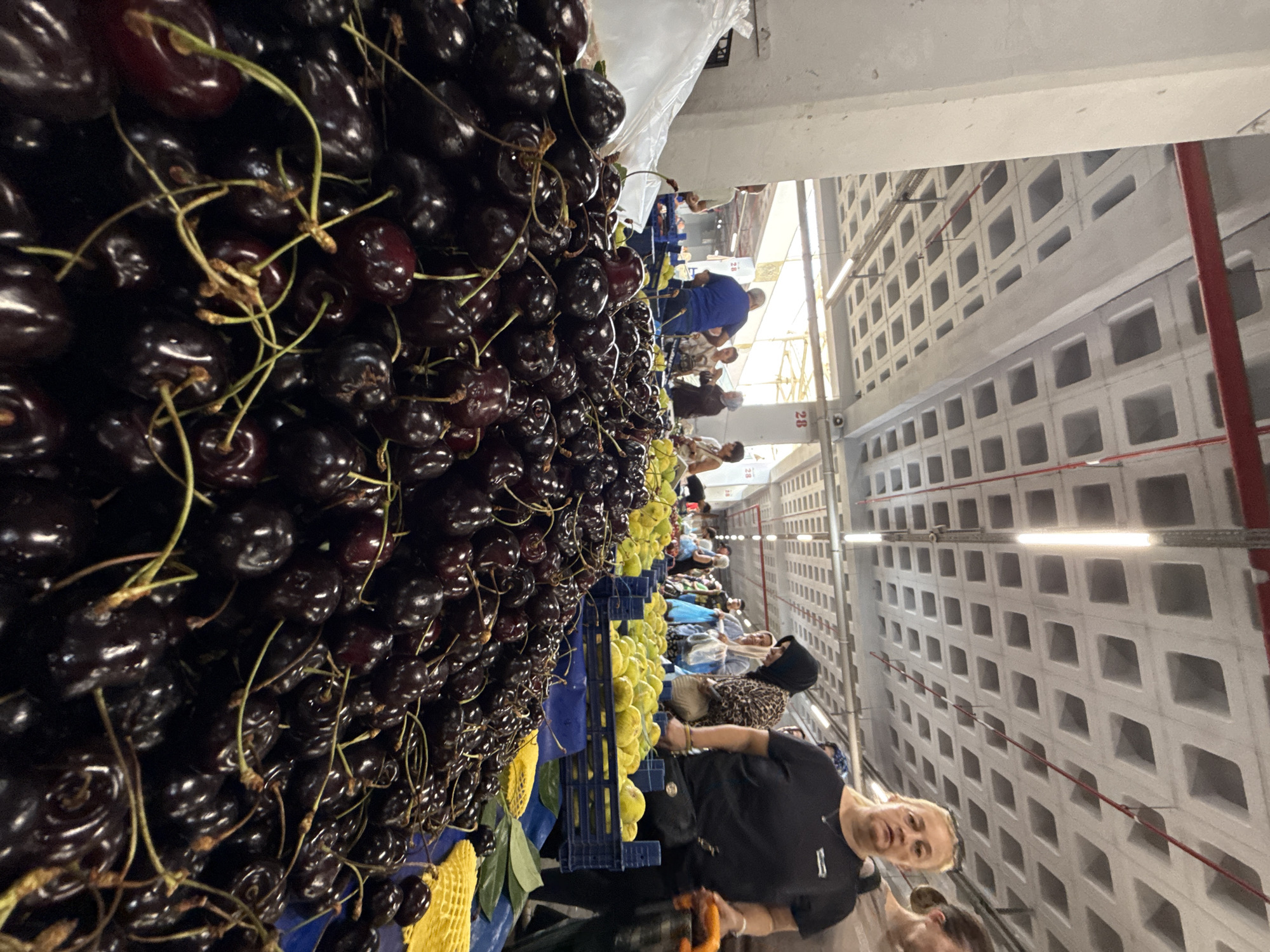 This is a vertical, eye-level shot of a market stall filled with dark red cherries. The foreground is dominated by a large pile of cherries with visible stems, obscuring the market table. In the midground, a woman with blonde hair and a black t-shirt is looking directly at the camera. Behind her, other shoppers and vendors are visible, some carrying shopping bags. In the background, a distinctive industrial ceiling with square openings and red pipes is prominent. The scene appears to be an indoor market, possibly the Kadıköy Market in Istanbul, Turkey, given the context of the city. The lighting suggests an indoor, possibly covered, market environment.