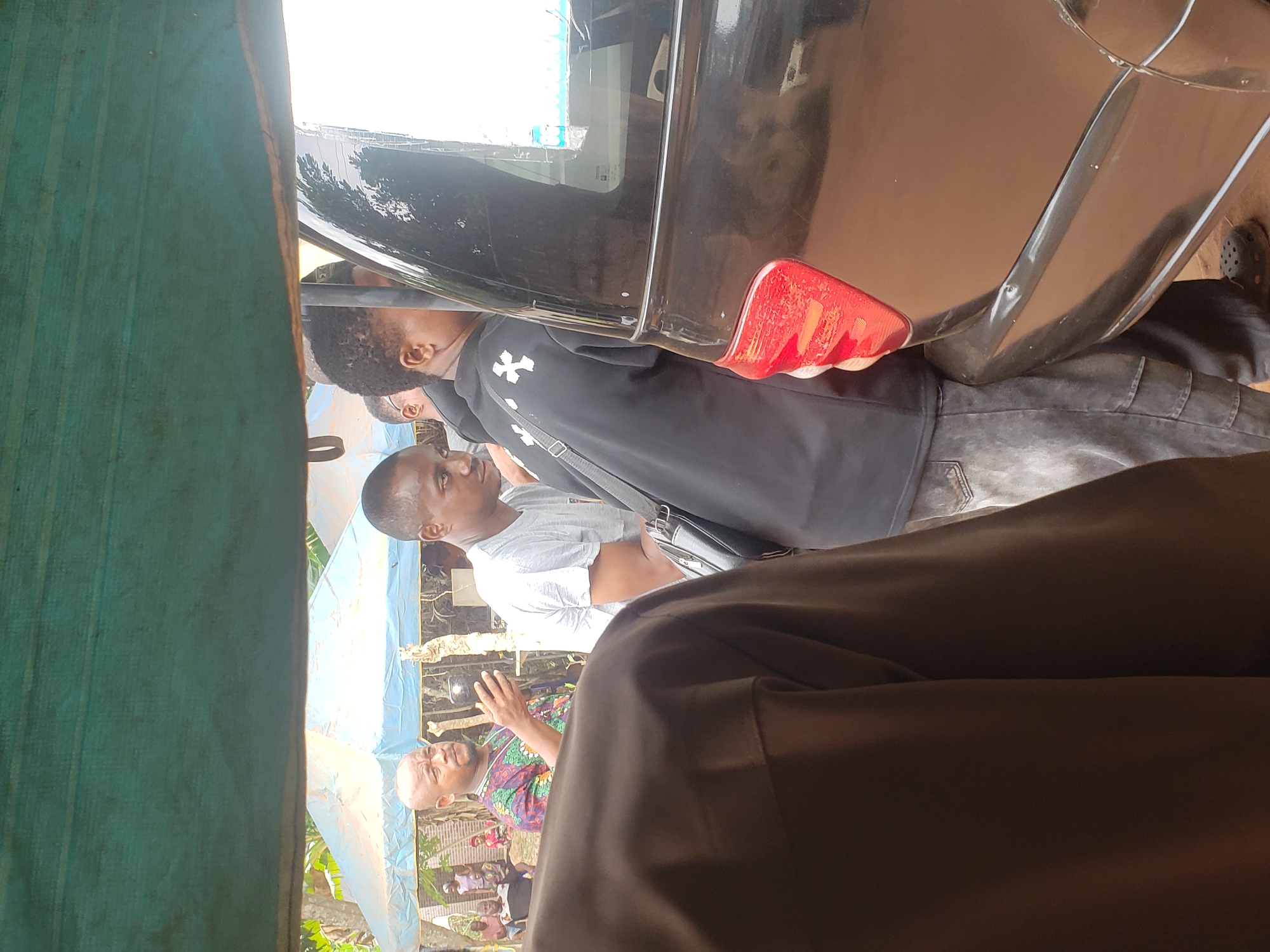 The image, taken in Umundugba, Nigeria, captures an outdoor gathering of several men during the daytime. The scene is framed by a green fabric awning on the left and the brown clothing of the person holding the camera in the right foreground.
In the mid-ground, a black car, likely a sedan or SUV, is parked, with its rear section, including a prominent red taillight and side windows, clearly visible. Two men are standing close to the car. One man, wearing a white short-sleeved t-shirt and a black shoulder bag, has short dark hair and faces towards the left of the frame. Behind him, another man in a black hoodie with a distinct white cross-like emblem on the chest and dark jeans is positioned near the car's rear passenger door. A person with a beard is partially visible near the front of the car, reflected in the window.
Further to the right, a bald man wearing a vibrant, multi-colored patterned shirt is actively gesturing with his right hand, suggesting he is speaking or explaining something to the group. Other individuals are faintly visible in the background, standing under what appears to be a light blue tarp or canopy, providing shade. The ground beneath them seems unpaved, consistent with an outdoor setting. The lighting is bright and natural, indicating it is daytime, possibly with an overcast sky given the lack of harsh shadows. The overall atmosphere suggests a casual community interaction or event. There is no discernible text visible within the frame.