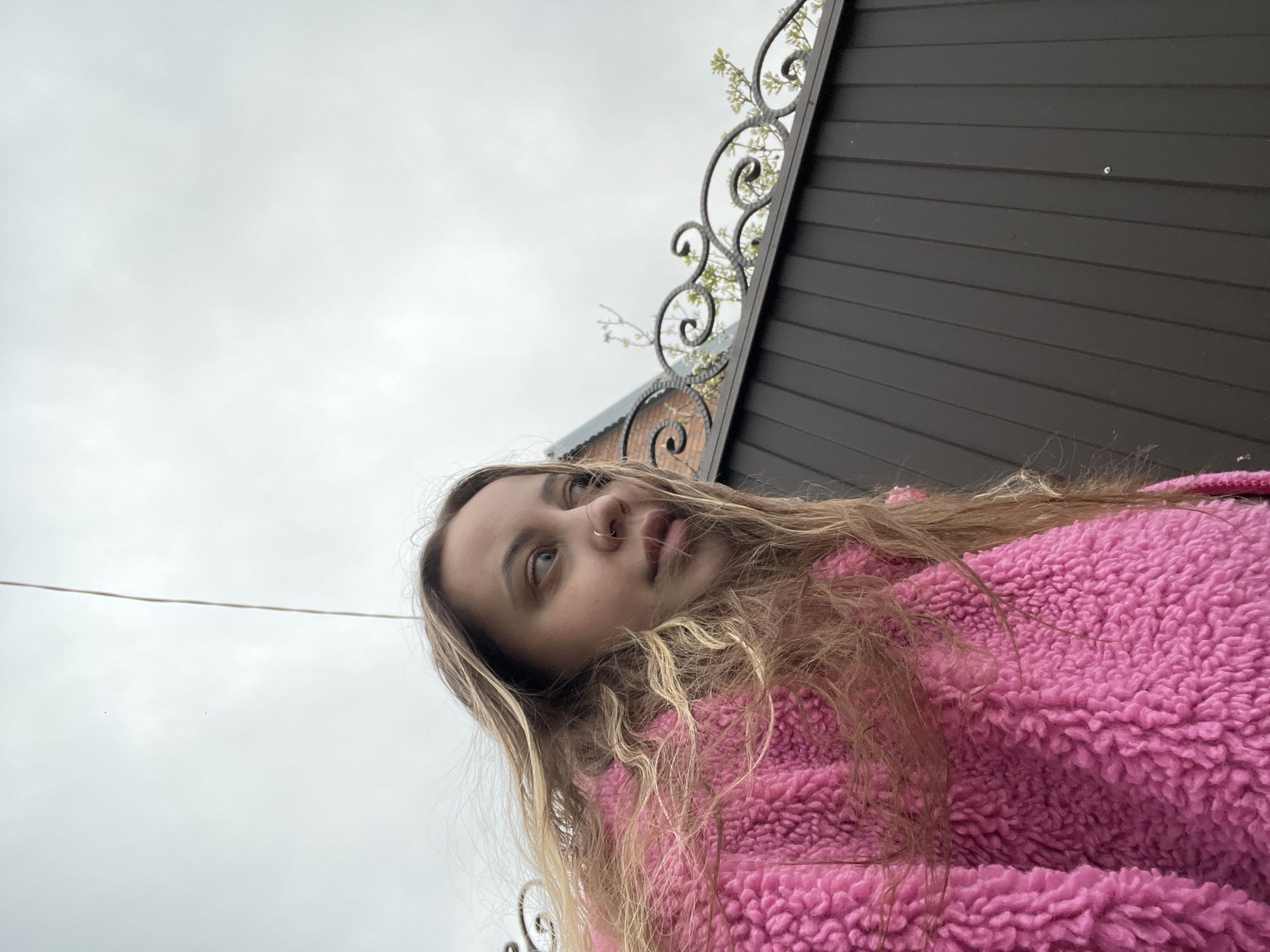 The image features a young woman wearing a pink, fluffy jacket, angled upwards towards the sky. The background includes a cloudy sky, a portion of a dark brown wooden fence, and a wrought iron decorative element. A building, possibly a house, is partially visible behind the fence. The location, Novopokrovskaya, Russia, is provided as context, but the image itself doesn't offer further specific details about the exact location within the city.