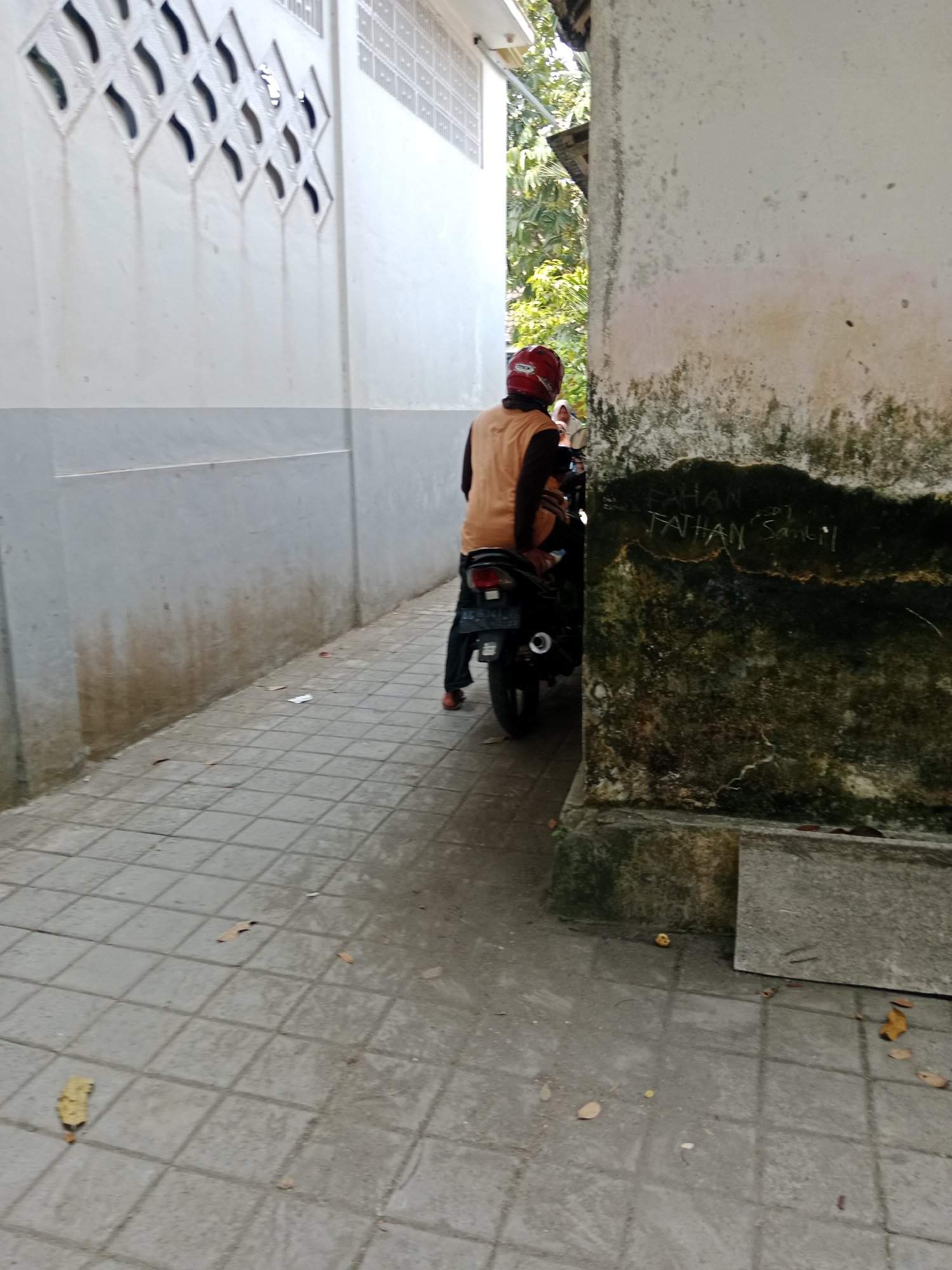 A medium shot captures a person wearing a red helmet and an orange vest while sitting on a dark-colored motorcycle. The motorcycle is positioned on a tiled pathway in a narrow passage between two buildings. The building to the left has a white wall with a grey horizontal stripe and decorative ventilation blocks. The building to the right has a weathered, off-white wall covered in patches of green moss and some faint graffiti. Leaves are scattered on the pathway. The scene is lit by daylight, casting shadows on the ground.