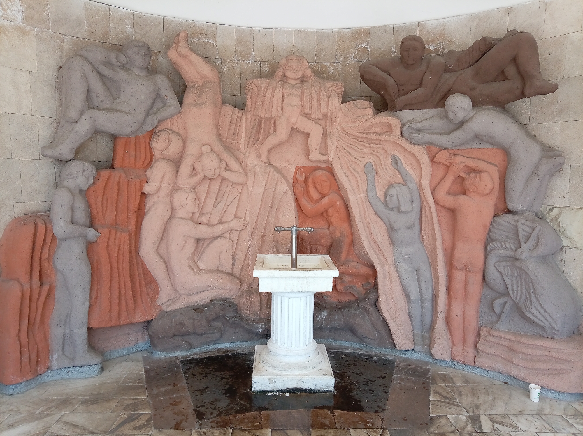 This image depicts a fountain in Zheleznovodsk, Russia, likely within a park or public space, given the surrounding architectural elements and the presence of a fountain. The fountain is integrated into a large, relief sculpture that features numerous stylized human figures in various poses, seemingly engaged in dynamic interactions or representing a narrative. The figures are rendered in a rough, textured style, with some figures appearing in a pinkish-orange hue and others in a gray or light beige. The sculpture itself is curved, suggesting it might be part of a wall or alcove.
At the center of the sculpture is the functional part of the fountain: a white, pedestal-mounted basin with a chrome spigot. The base of the fountain and the surrounding area appear wet, indicating recent or ongoing water flow. The floor in front of the fountain is tiled with a pattern of light and dark marble-like tiles, some of which are wet and stained. The wall behind the sculpture is constructed of light-colored, rectangular stone blocks.
The human figures in the relief sculpture are diverse in their depiction. Some are shown reclined, others are in active stances, and some appear to be supporting or reaching for others. There is a sense of movement and interconnectedness among the figures. The overall impression is of a monumental art installation designed to be both decorative and functional. A small, white paper cup is visible on the floor to the right of the sculpture, suggesting the presence of visitors.