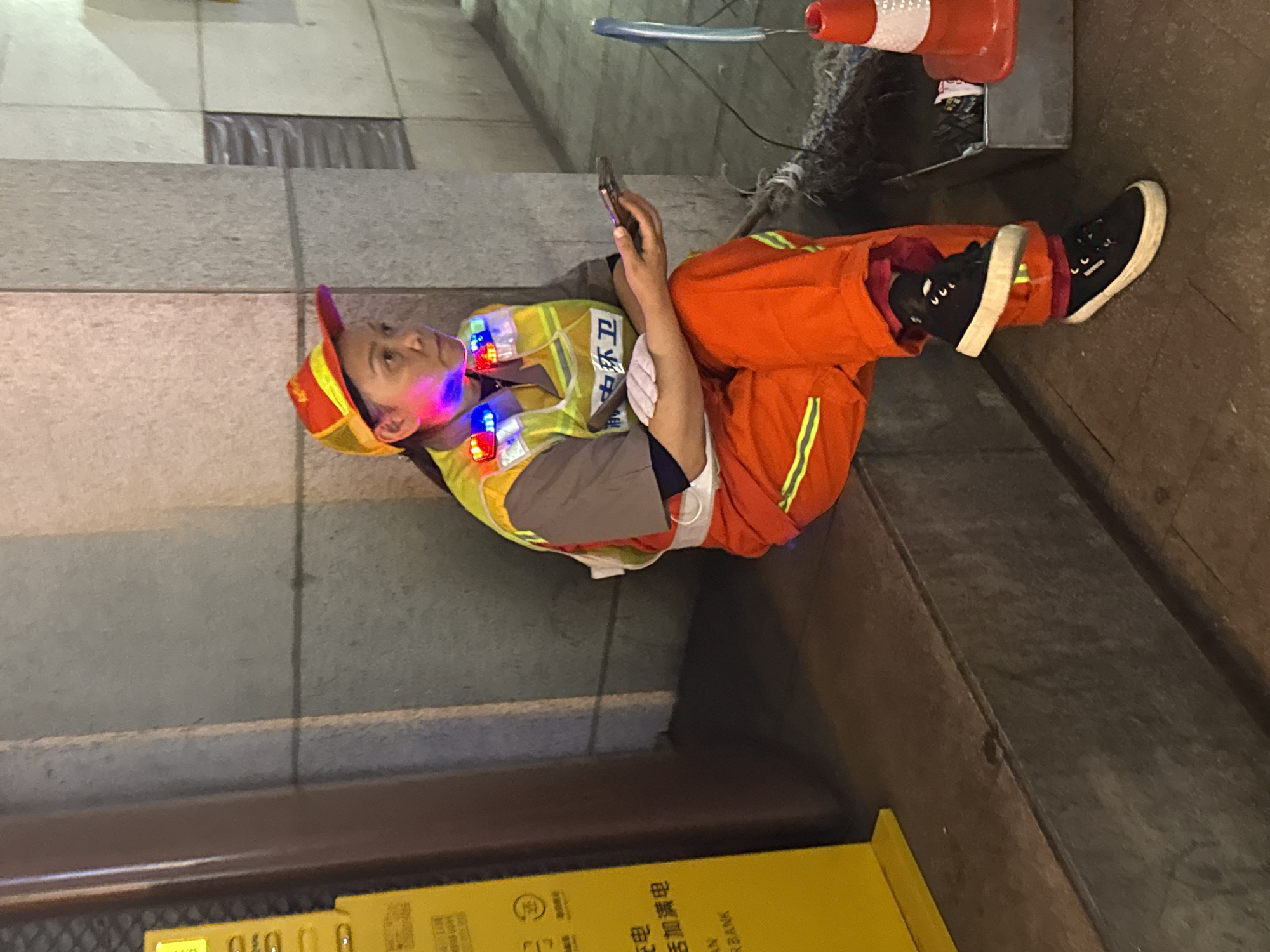 A person in a fluorescent yellow and orange uniform and a cap lies on a sidewalk and leans against a concrete wall, looking at a mobile phone. The person wears sneakers and has colorful lights attached to their clothing. Near the person is an orange traffic cone, a charging station, and some wiring. The uniform includes Chinese writing indicating "Environmental Sanitation". This scene is set in the city of Yuzhong District, China.
