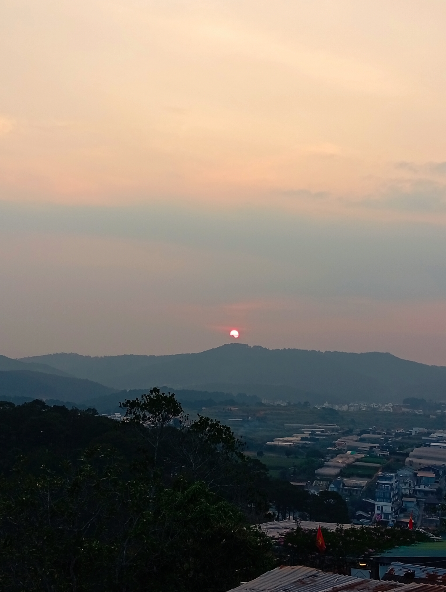 The image depicts a sunset over Da Lat, Vietnam, with a view of rolling hills in the mid-ground. The sun is partially obscured behind one of the hills, casting a warm glow in the sky. The foreground shows a mix of dense foliage and rooftops. The distant landscape includes rows of greenhouses, suggesting agricultural activity. Flags are visible in the lower right, indicative of Vietnam. The atmosphere is hazy, contributing to the muted color palette of the scene. Buildings, possibly residential, are sparsely scattered amongst the greenhouses. The overall setting appears rural or peri-urban.