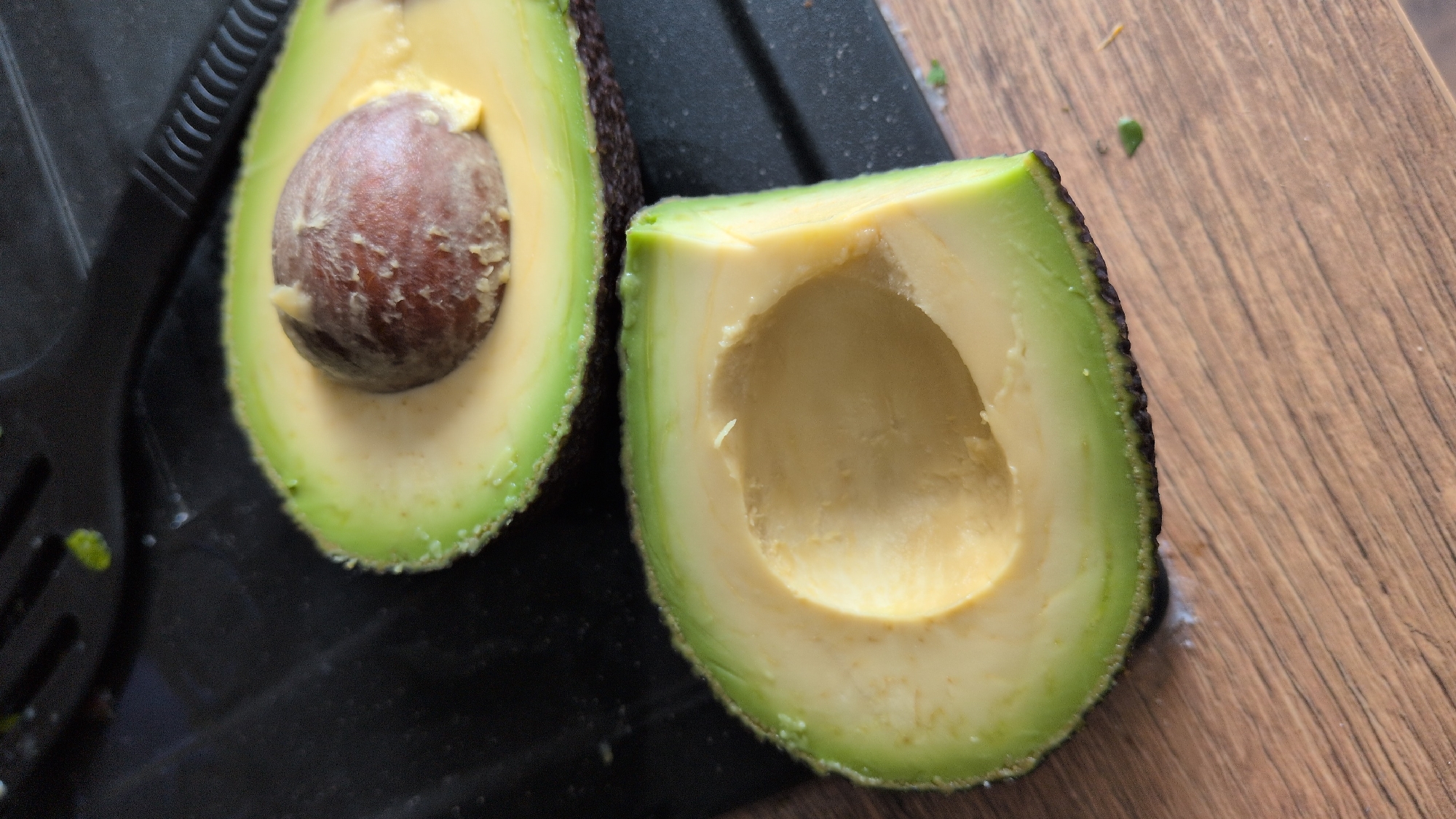 The image displays two halves of a ripe avocado on a wooden surface, with a black cutting board or platter visible on the left. One half of the avocado retains its large, brown pit, while the other half is hollowed out, showing the creamy texture of the fruit. The avocado's skin is dark green, transitioning to a vibrant green just beneath the skin, and then to the pale yellow flesh. A black utensil, possibly a fork or spatula, is partially visible on the left, adjacent to the avocado half with the pit. The wooden surface has a visible grain pattern. The lighting suggests an indoor setting, possibly a kitchen, with no specific time of day or weather conditions indicated. There is no visible text in the image.