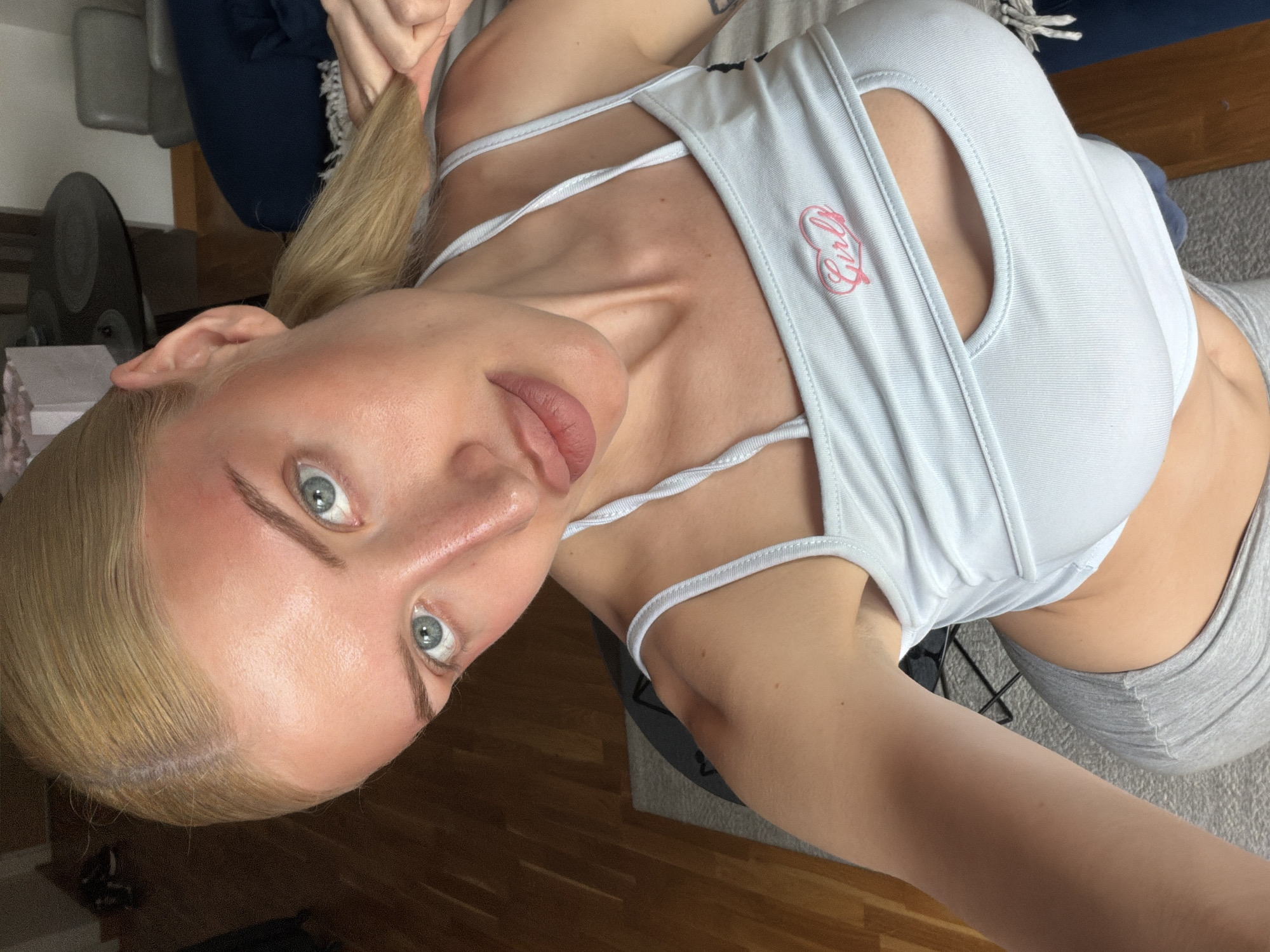 A fair-skinned woman with blonde hair and light blue eyes is the primary subject of this selfie-style photograph. She is wearing a light blue sports bra with thin straps and a "Girl" logo in pink lettering. The top features a keyhole neckline. She is also wearing light grey pants.
The background reveals elements of an interior setting. A dark blue upholstered chair and a black fan are visible, indicating a domestic environment. The floor is wooden.
The geographical location of Belgrade, Serbia, cannot be determined from this image. There are no definitive visual cues present to confirm that location.
