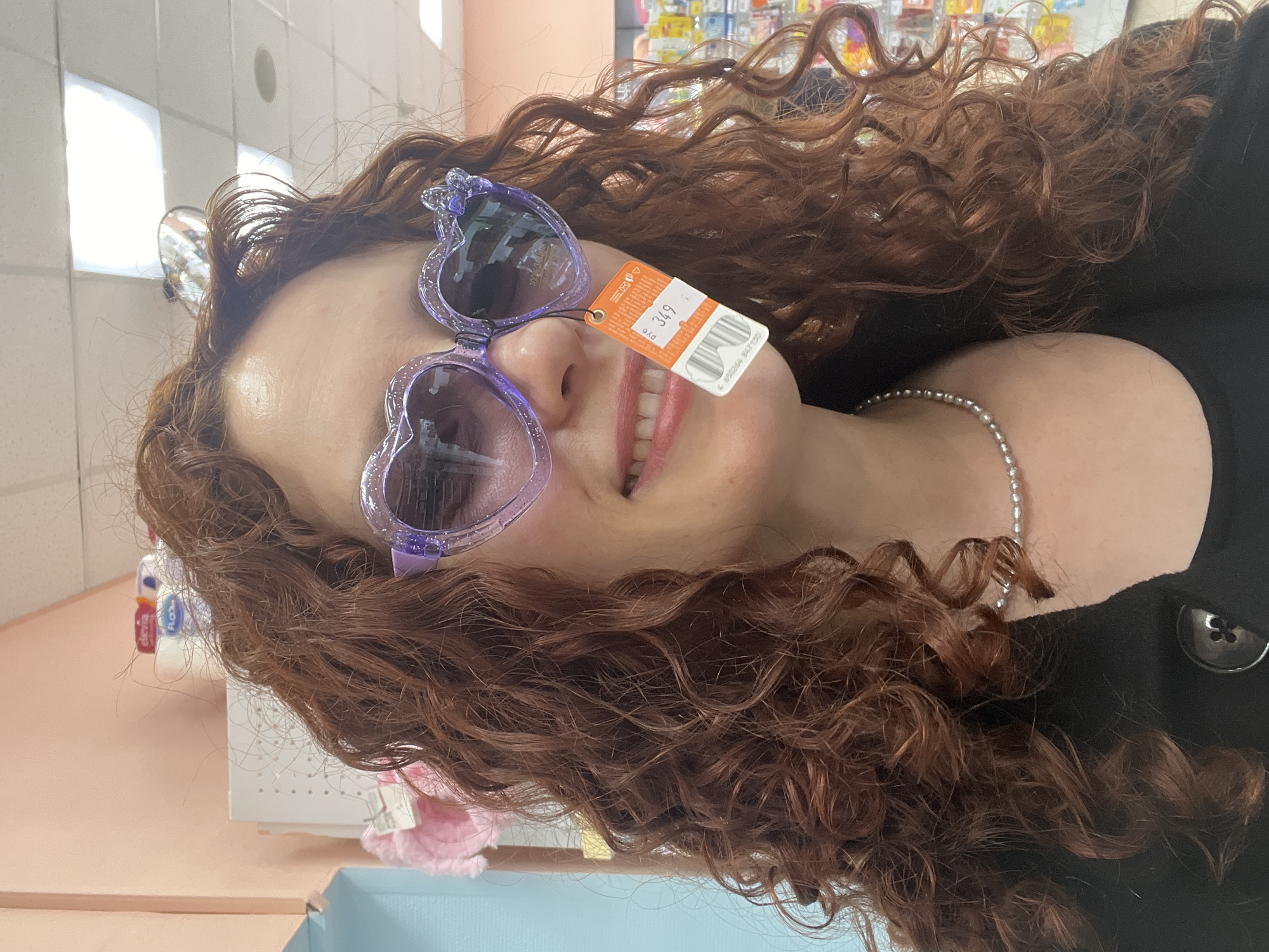 A woman with curly red hair is wearing purple heart-shaped sunglasses with a price tag hanging from them. She is smiling slightly. She is wearing a black top and a beaded necklace. The background appears to be a store, possibly a retail or pharmacy setting, with shelves stocked with merchandise. Based on the background, it is difficult to confirm the location is in Saint Petersburg, Russia.