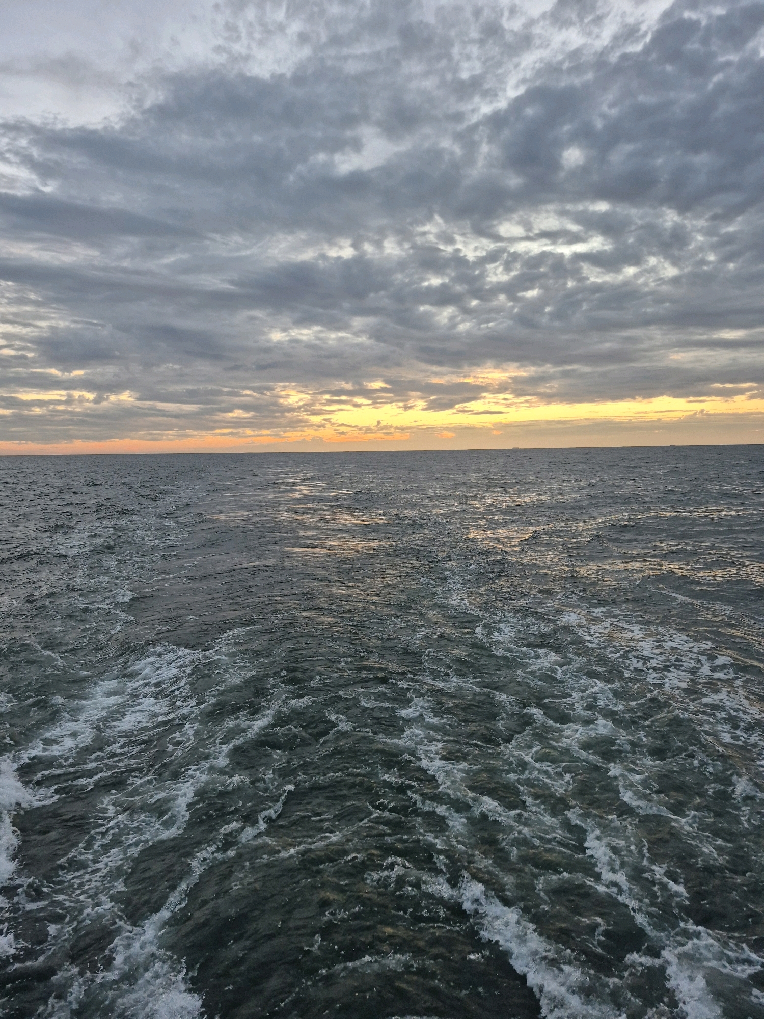 The image shows a dark, choppy sea with a white wake extending from the bottom of the frame towards the horizon. The sky above is overcast with gray clouds, but there is a band of pale yellow and orange light near the horizon, suggesting either sunrise or sunset. The overall scene is one of open water under a cloudy sky. There are no discernible features of a city in Germany or any people or specific objects.
