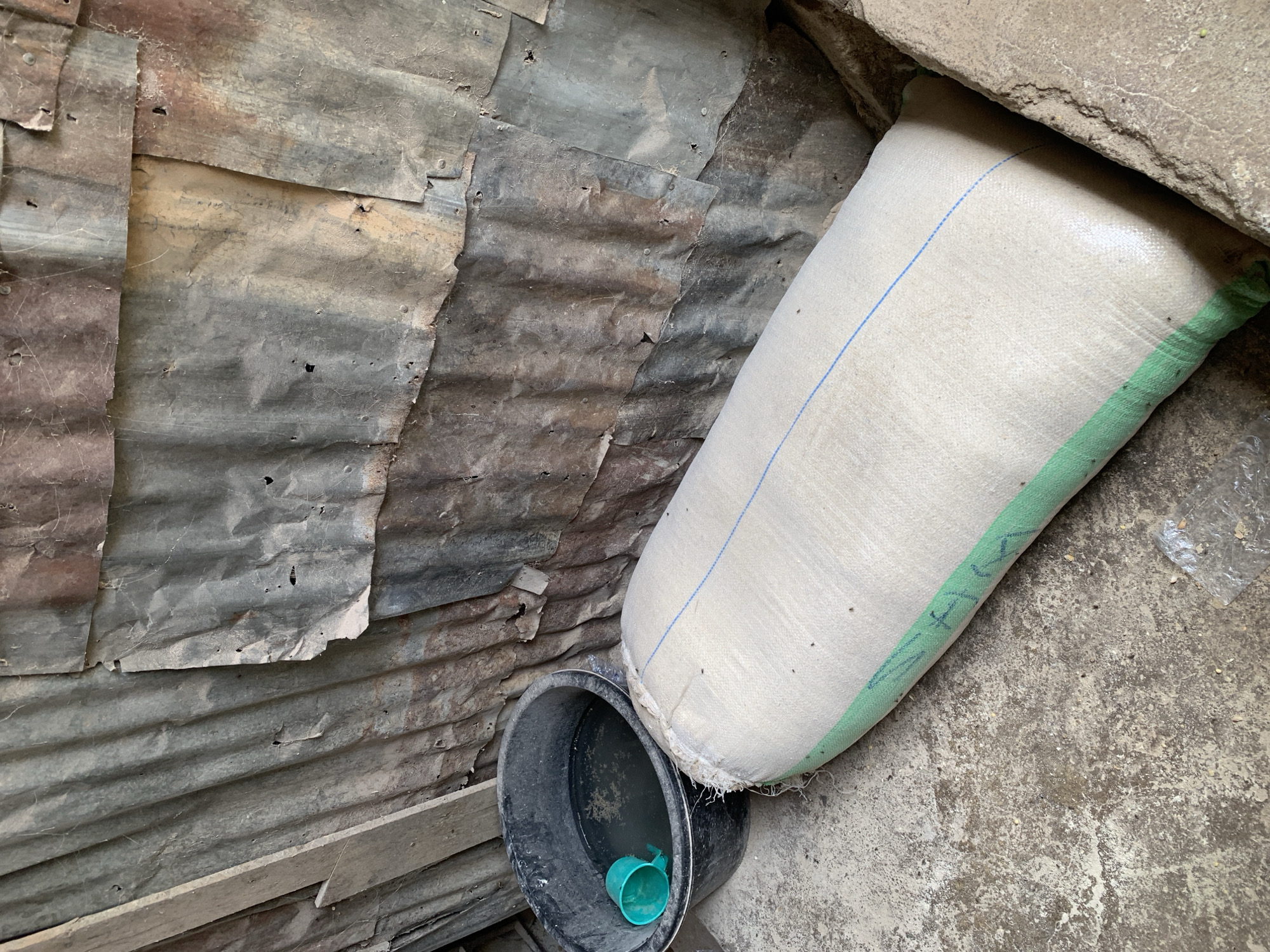 The image shows an interior space in Unknown, Unknown. The left wall is constructed of weathered corrugated metal sheets attached with rivets or nails. In the foreground is a black bucket with some water inside, and a small green plastic cup is partially submerged in the water. Next to the bucket is a large, off-white woven bag with green and blue stripes leaning against a concrete wall. The floor is also concrete. The scene appears to be a storage area or a simple shelter. There are some debris on the floor.