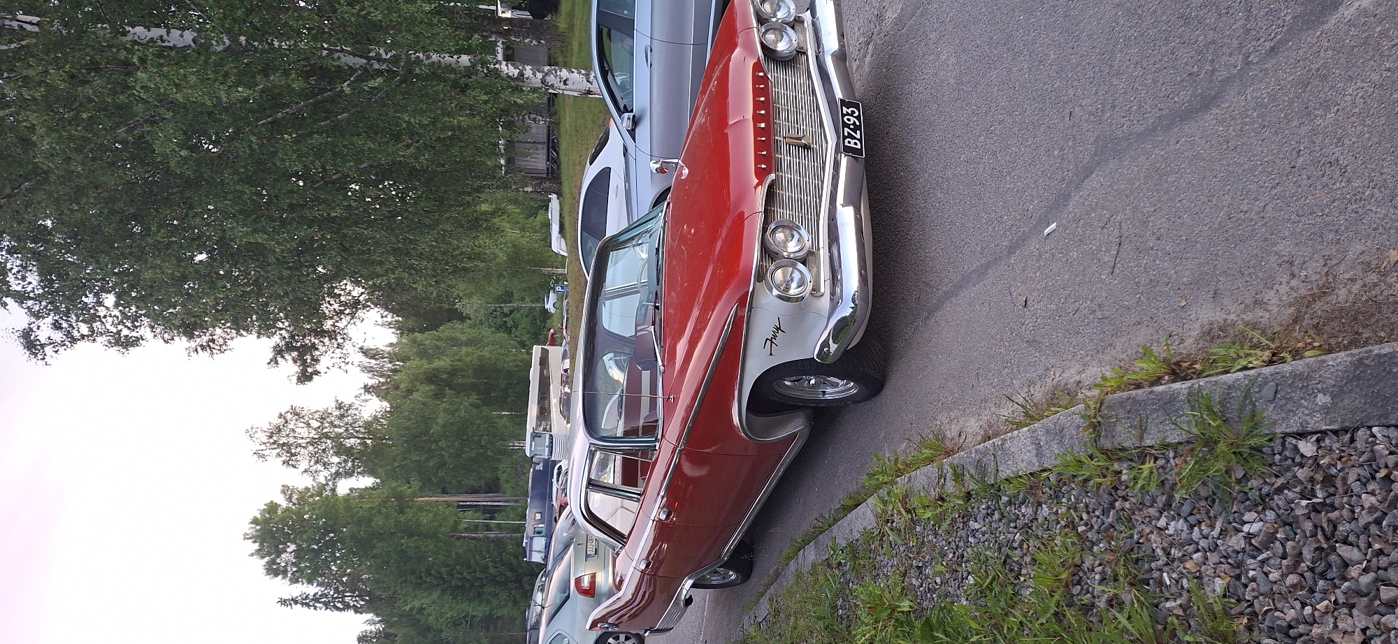 In Salpakangas, Finland, a vintage red and white Plymouth Fury sedan is parked on the right side of a paved road, adjacent to a gravel and grass verge. The car, which has chrome accents and a distinctive grill, faces towards the left side of the frame. In the background, a line of trees, including birches and other deciduous trees, creates a backdrop. Behind the trees, various vehicles are visible, including a white bus and a silver car, suggesting a parking lot or a road with other parked vehicles. The sky is a pale, overcast hue.