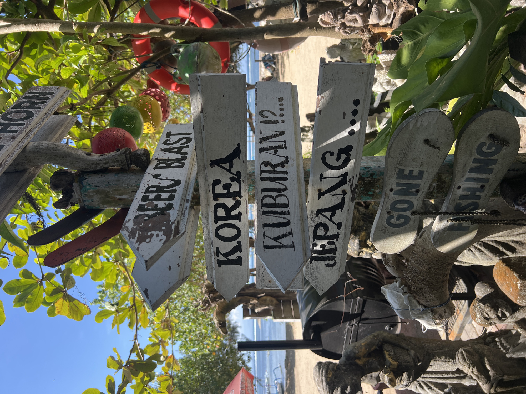 A weathered wooden signpost with multiple directional arrows stands on a beach in Denpasar, Indonesia. The signs are painted white with black lettering and point in various directions. Some of the destinations indicated are "BEER BLAST," "KOREA," "KUBUAN !? ...," "JEPANG ...," "GONE," and "FISHING." Behind the signpost, lush green foliage and the bright blue sky are visible, along with the ocean and a beach. Various decorative objects, including colorful spheres and a red life preserver, are hanging from the signpost and nearby trees. In the foreground, several stone sculptures are present. A black barbecue grill is also visible behind the signpost. The overall scene suggests a casual, tropical atmosphere.