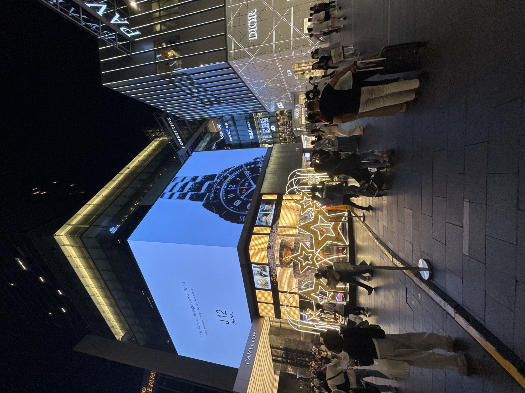 The image captures a street scene at night in Kuala Lumpur, Malaysia. The shot is taken from a low angle, highlighting the tall buildings and illuminated storefronts. On the left, a large digital billboard displays a Chanel J12 watch advertisement, alongside the text "In The Greatest Strength Lies Softness." Next to the digital billboard is a building with the word "PAVILION" vertically lit, further identifying the location. Adjacent to this, storefronts feature illuminated star decorations. On the right side of the frame is a Dior store with intricate geometric lighting patterns. Pedestrians are walking along the tiled pavement. Some are pulling luggage. Other storefronts are visible, including those of Toys "R" Us and Muji. The environment suggests a high-end retail district, bustling with activity.