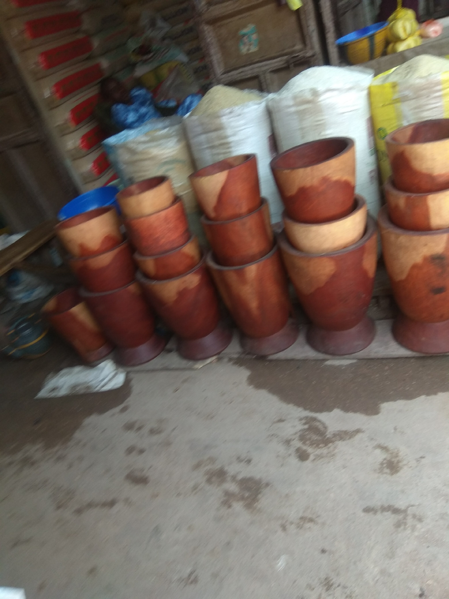 The image depicts a market stall in Jalingo, Nigeria, displaying a collection of wooden mortars and pestles for sale, alongside large sacks of what appears to be grain or flour. The mortars are of various sizes and are stacked or arranged in rows, with their characteristic wooden texture and reddish-brown coloration visible. Some of the mortars are nested inside each other. Behind the mortars, large white and yellow sacks are stacked, suggesting a bulk commodity sale. A blue plastic bowl is visible on the left side of the frame. The ground beneath the items appears to be a paved surface, with some wet patches, indicating possible recent rain or water spillage. The overall lighting suggests an outdoor or semi-covered market environment, with a sense of everyday commerce.