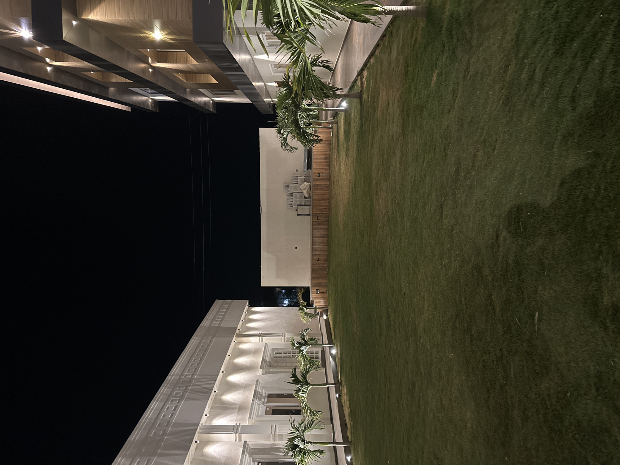 The image shows an outdoor view at night in Kalamb, India. Two buildings stand opposite each other with a stretch of green lawn in between. Palm trees are planted along the edges of the lawn on both sides. The building on the left is white and illuminated with multiple lights. The building on the right has a dark wood or stone-like facade on top, with lights accentuating its structure. The sky is black and empty.