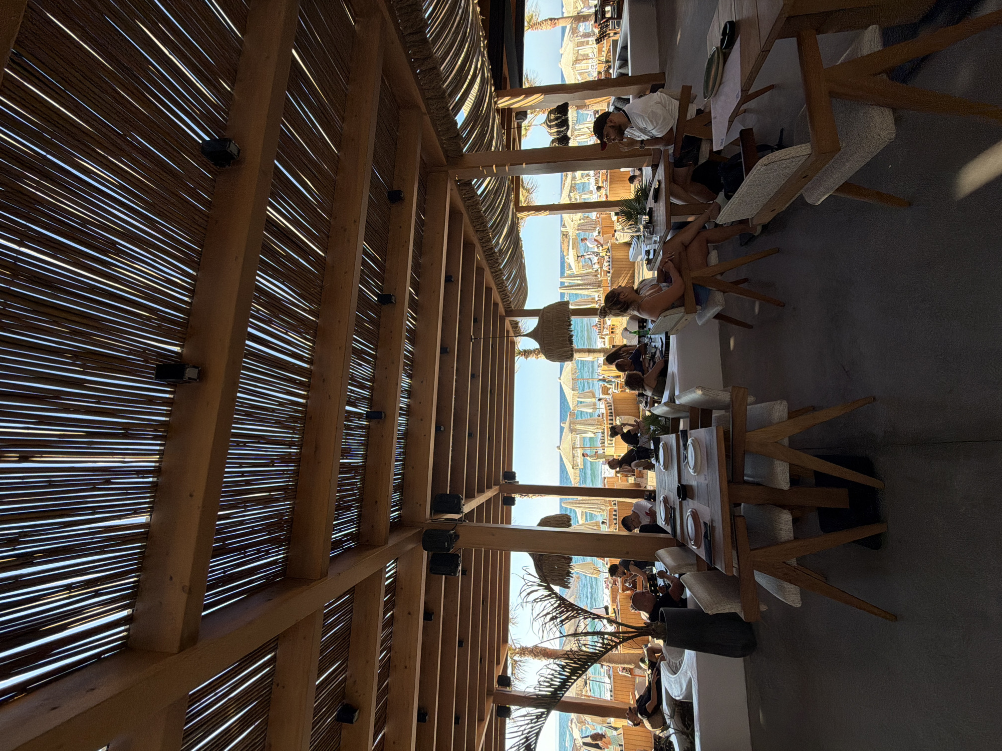 This image shows the interior of a beachside restaurant in Malia Municipal Unit, Greece, with a view of the sea and beach in the background. The restaurant features a rustic, open-air design with a thatched roof supported by wooden beams. Several people are seated at tables, some dining, others engaged in conversation. In the foreground, a wooden table is set with plates and cutlery. Beyond the seating area, beach umbrellas and lounge chairs are visible on the sandy beach, with the turquoise sea stretching to the horizon under a clear blue sky. The overall atmosphere appears relaxed and casual, typical of a seaside establishment.