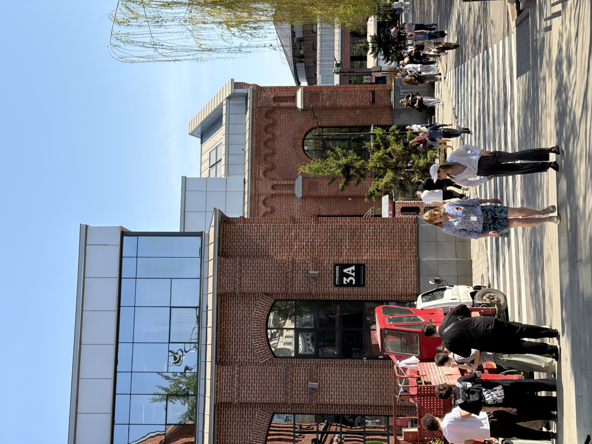 The image shows an outdoor scene in Moscow, Russia. A group of people is walking across a pedestrian crosswalk near a building with brick and glass facades. A red utility vehicle is parked near the building. A sign on the building reads "СТРОЕНИЕ 3А." The environment is sunny with long shadows cast by the people and buildings. The presence of multiple people suggests a public space or gathering area.