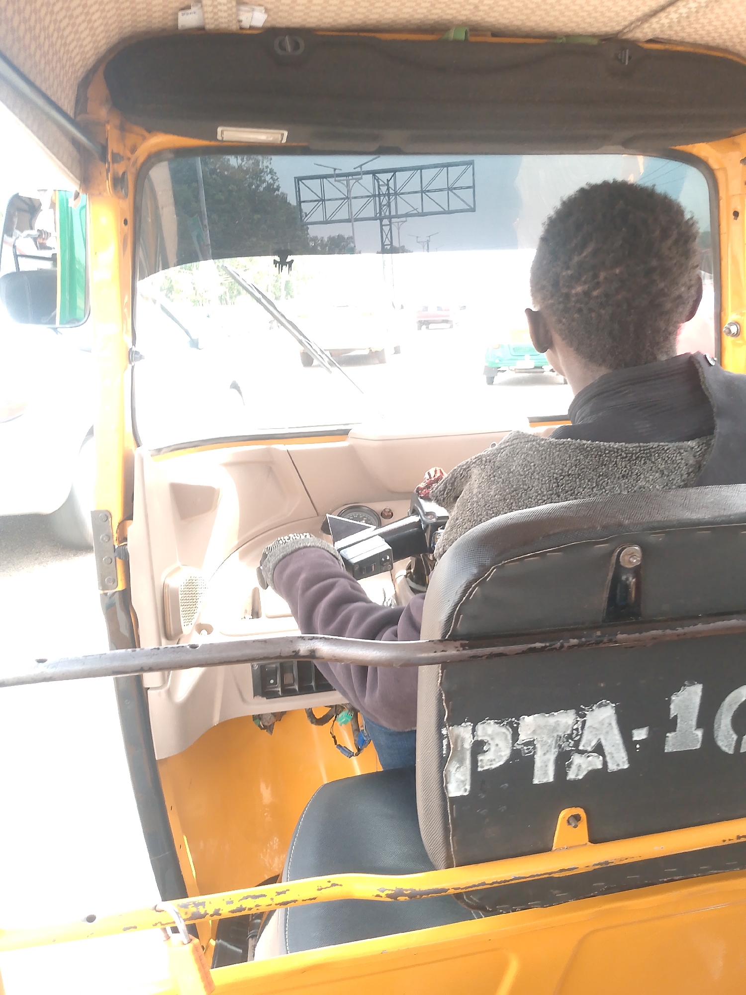 The image shows the interior of a yellow tuk-tuk, a three-wheeled vehicle, in Bukuru, Nigeria. The driver, an individual with short, dark hair, is visible from the back, operating the vehicle. They are wearing a dark jacket over a grey shawl. A black seat with "PTA-10" written on the back is visible in the foreground. The windshield shows a street with other vehicles and an overhead structure. The tuk-tuk's interior is visible, including the dashboard and steering mechanism. Sunlight is apparent.