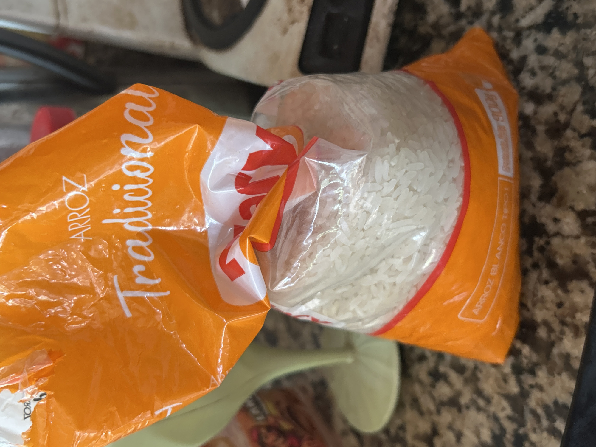 The image shows a bag of rice on a speckled countertop. The bag is mostly orange with white lettering that reads "ARROZ Tradicional". The lower part of the bag is clear plastic, revealing white rice. There is also text at the bottom of the bag that appears to say "ARROZ BLANCO TIPO I" and "Contenido Net: 900g". In the background, a light green object, possibly a pitcher or vase, is visible to the left. To the right, a kitchen appliance, possibly a rice cooker or blender, is partially visible. The overall scene appears to be indoors, likely in a kitchen. There are no people or actions depicted in the image. The image is a close-up of the bag of rice, with the background slightly out of focus.