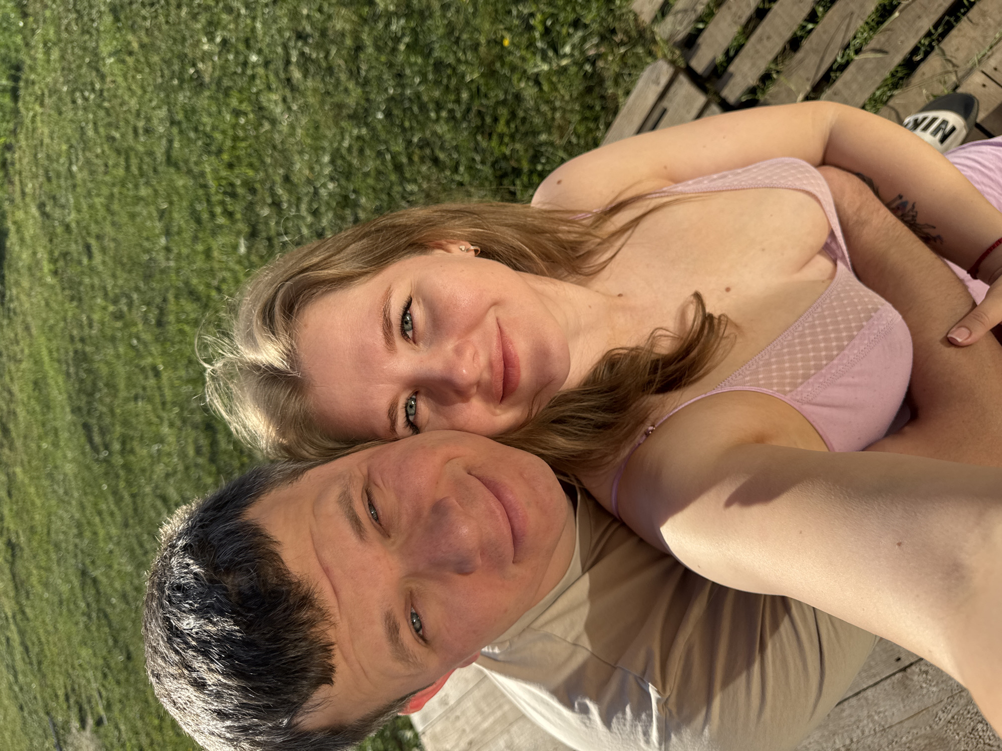 A selfie depicts a man and a woman lying down in an embrace. The woman is wearing a light pink top and the man is wearing a light brown t-shirt. A small portion of a wooden plank structure is visible in the upper right and lower left corners of the frame. Grass is visible surrounding the subjects. The photo's location is likely Ферма, Russia.