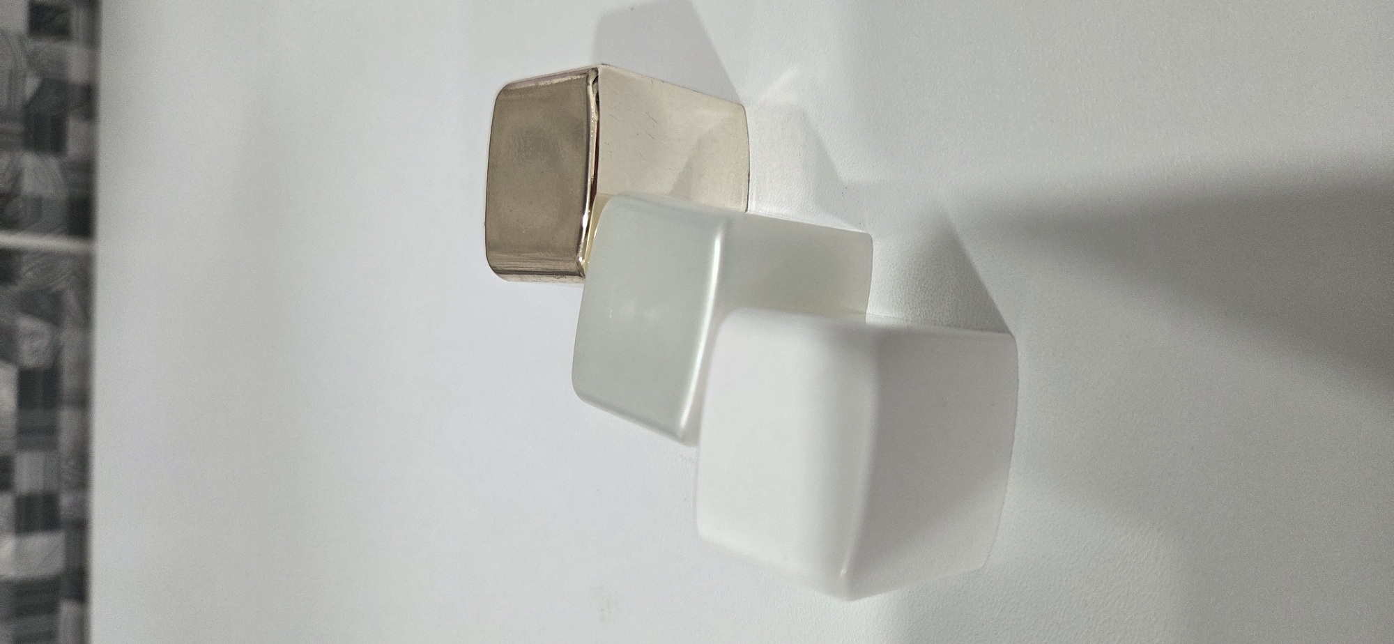 The scene, located in the city of Lipetsk, Russia, displays three rectangular objects with rounded corners positioned diagonally on a flat, light-colored, subtly textured surface. The object in the foreground, at the bottom-left, is opaque white. Overlapping it to the right and slightly behind is a translucent object with a faint green or aqua tint. Further to the right and behind the translucent object is a reflective, metallic object, appearing gold or brass in color. Shadows are cast by the objects onto the surface. The background features a blurry pattern of dark gray and white squares or tiles.