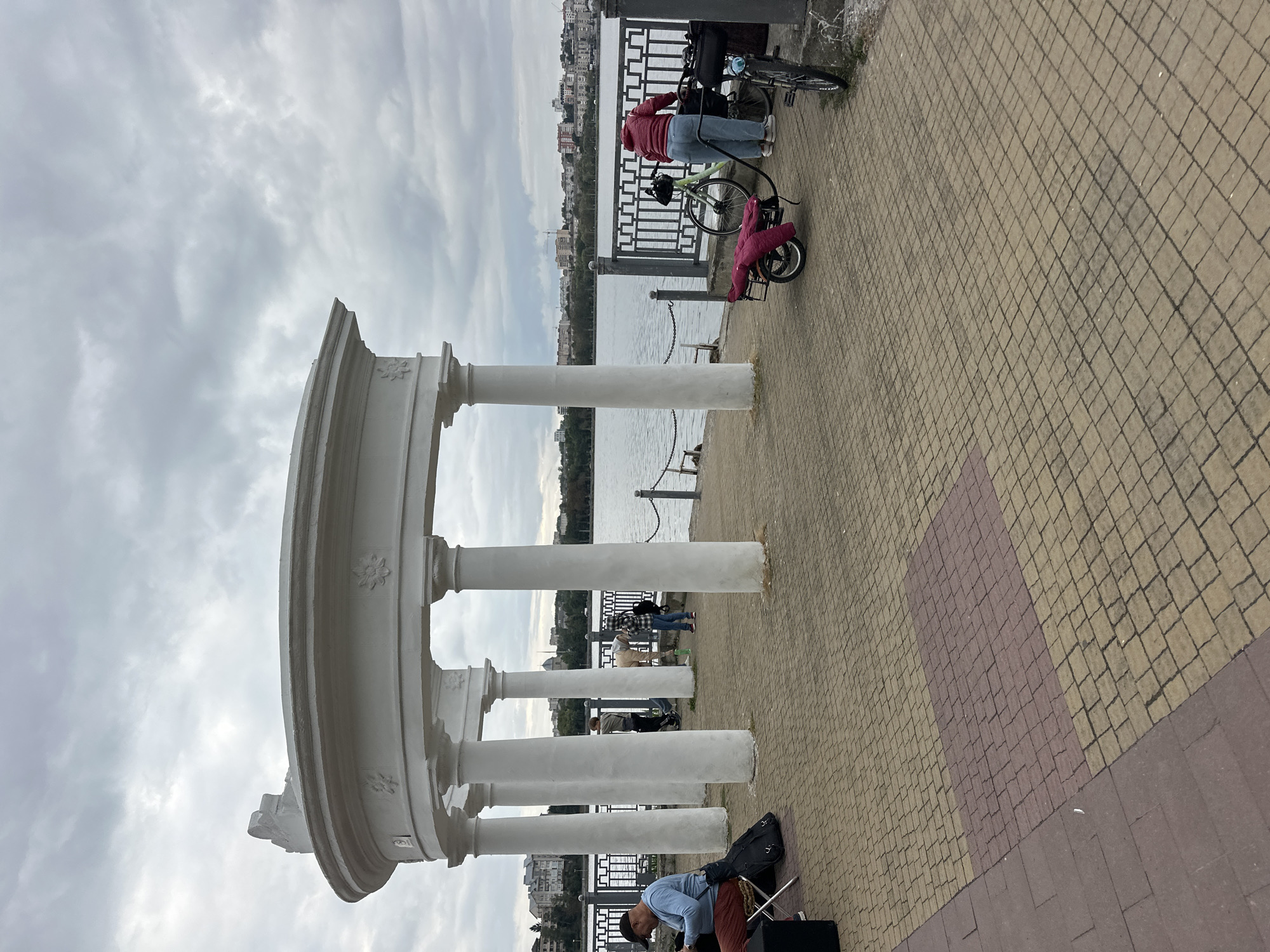 The image depicts an outdoor scene by a body of water, likely Ternopil Lake in Ternopil, Ukraine, given the architectural style. In the foreground, a paved walkway with a pattern of yellow and red bricks leads towards a white, open-air rotunda supported by classical columns. A man in a blue shirt and dark pants sits on a chair, looking down at something in his hands, with a black guitar case next to him. Further back, near the rotunda, several people are visible. One person is bent over, possibly attending to a bicycle with a trailer attached. Another person is standing, and a third person is partially obscured. A metal railing separates the walkway from the water. Across the water, a cityscape with buildings and trees is visible under a cloudy sky. The overall atmosphere suggests a calm, overcast day in a public park or promenade.