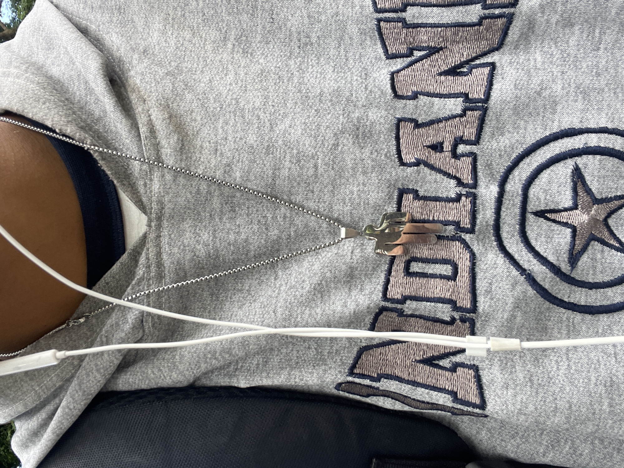 The image shows a close-up of a person wearing a grey hoodie with embroidered lettering that appears to read "INDIANAPOLIS". A silver chain necklace with a geometric pendant is visible around the neck, and white earbuds are also present. The person is also wearing a black backpack strap over their left shoulder. The background is blurred but suggests an outdoor setting with some greenery. There is no specific information available about the location in El Valle del Espíritu Santo, Venezuela, as the image provides no contextual clues.