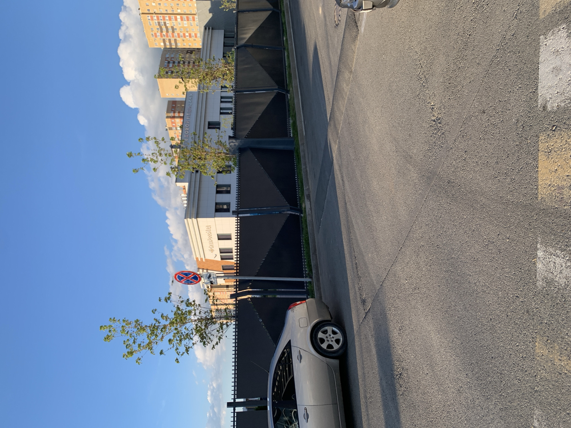 The image shows a street scene with buildings and a car. The buildings are modern, with light-colored facades and many windows. One building has Cyrillic text on it that reads "КЛИНИЧЕСКИЙ МЕДИЦИНСКИЙ ЦЕНТР". There is a black fence in front of the buildings, and a tree with green leaves is growing nearby. A silver car is parked on the side of the asphalt road, which has yellow and white markings. A traffic sign with a red circle and a blue cross inside indicates no stopping. The sky is blue with some white clouds. The lighting suggests it is daytime, likely late afternoon due to the long shadows. The scene appears to be in an urban environment in Stavropol, Russia. There are no people visible in the image.